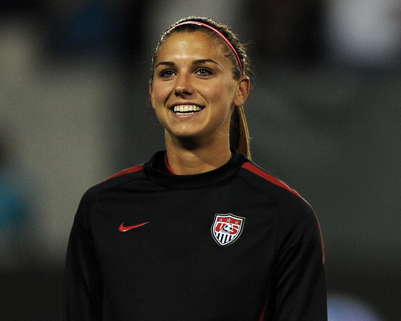 Alex Morgan 桌布 壁紙 — 一位微笑的女足球員，身穿帶有紅色點綴和粉紅色頭帶的黑色美國國家隊訓練球衣。