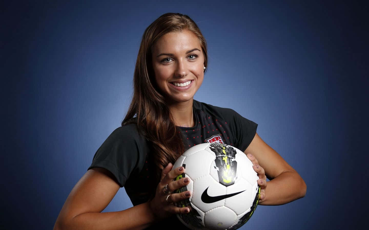 Alex Morgan 桌布 壁紙 — 一位微笑的女足球員在深藍色攝影棚背景前，手持一個白色的Nike足球。