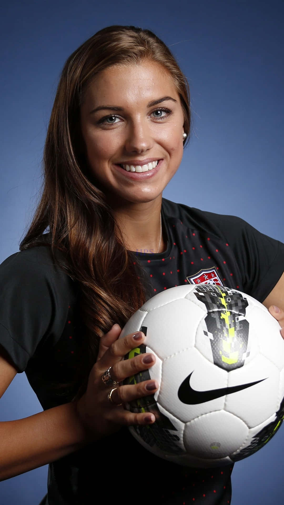 Alex Morgan 桌布 壁紙 — 一位微笑的女足球員身穿黑色球衣，在純藍色攝影棚背景前拿著一個白色Nike足球。