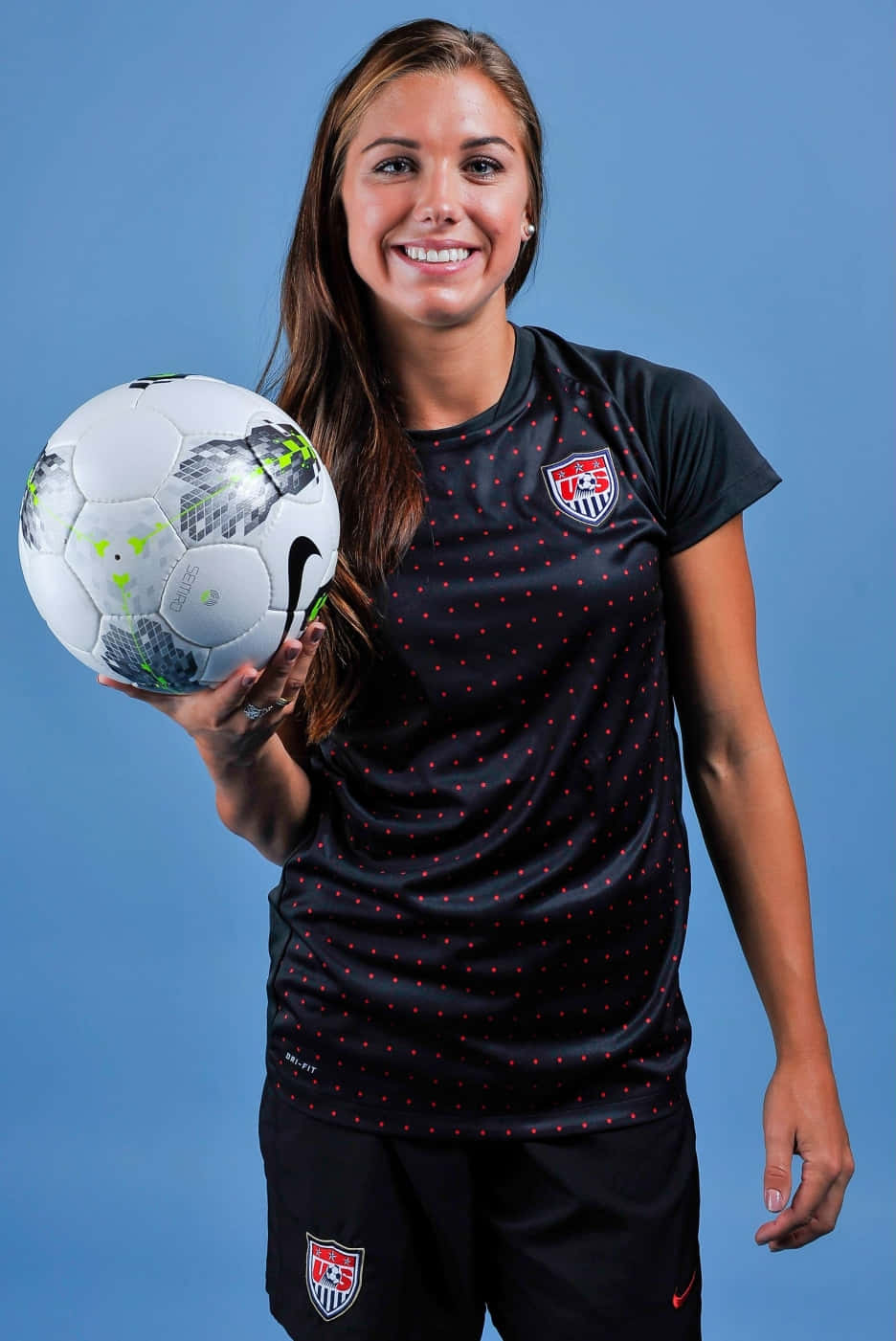 Alex Morgan 桌布 壁紙 — 一位微笑的女足球員身穿黑色美國國家隊球衣，在純藍色攝影棚背景前抱著一個白色足球。