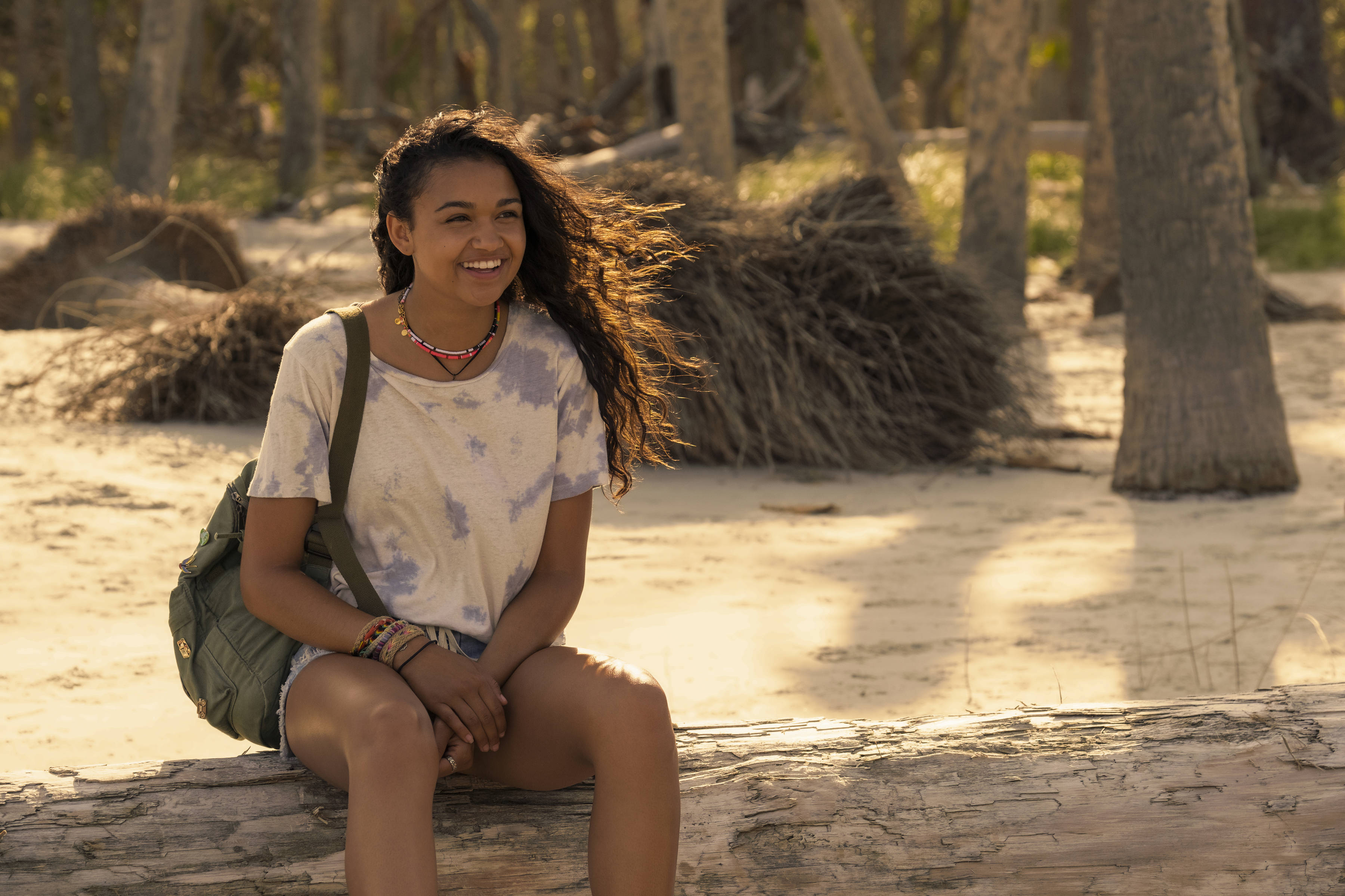 《Outer Banks》Madison Bailey 壁紙 — 一位微笑的年輕女子，頭髮被風吹亂，坐在陽光普照的沙地森林中的一根木頭上，穿著紮染襯衫，背著綠色背包。