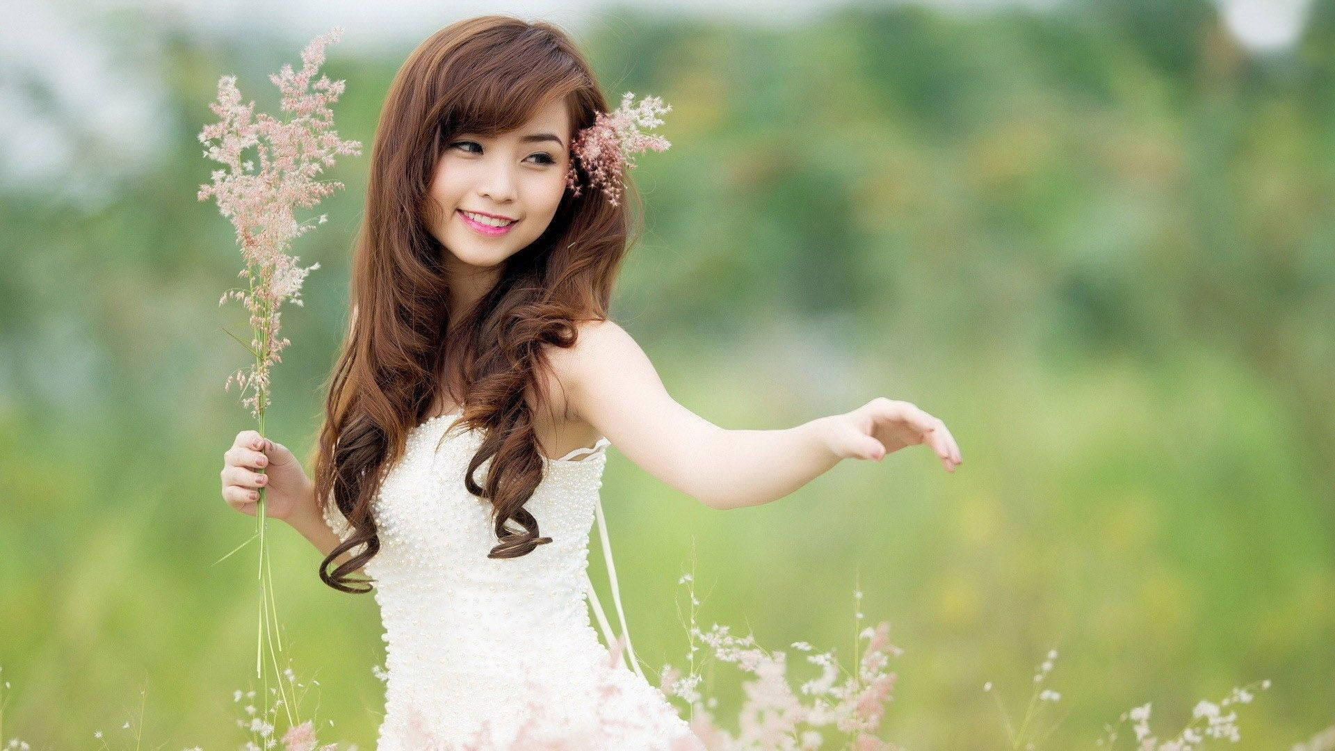 Cute Girl 桌布 壁紙 — 一位微笑的年輕女子穿著白色連衣裙，在陽光明媚的綠色田野中手持粉紅色的草。