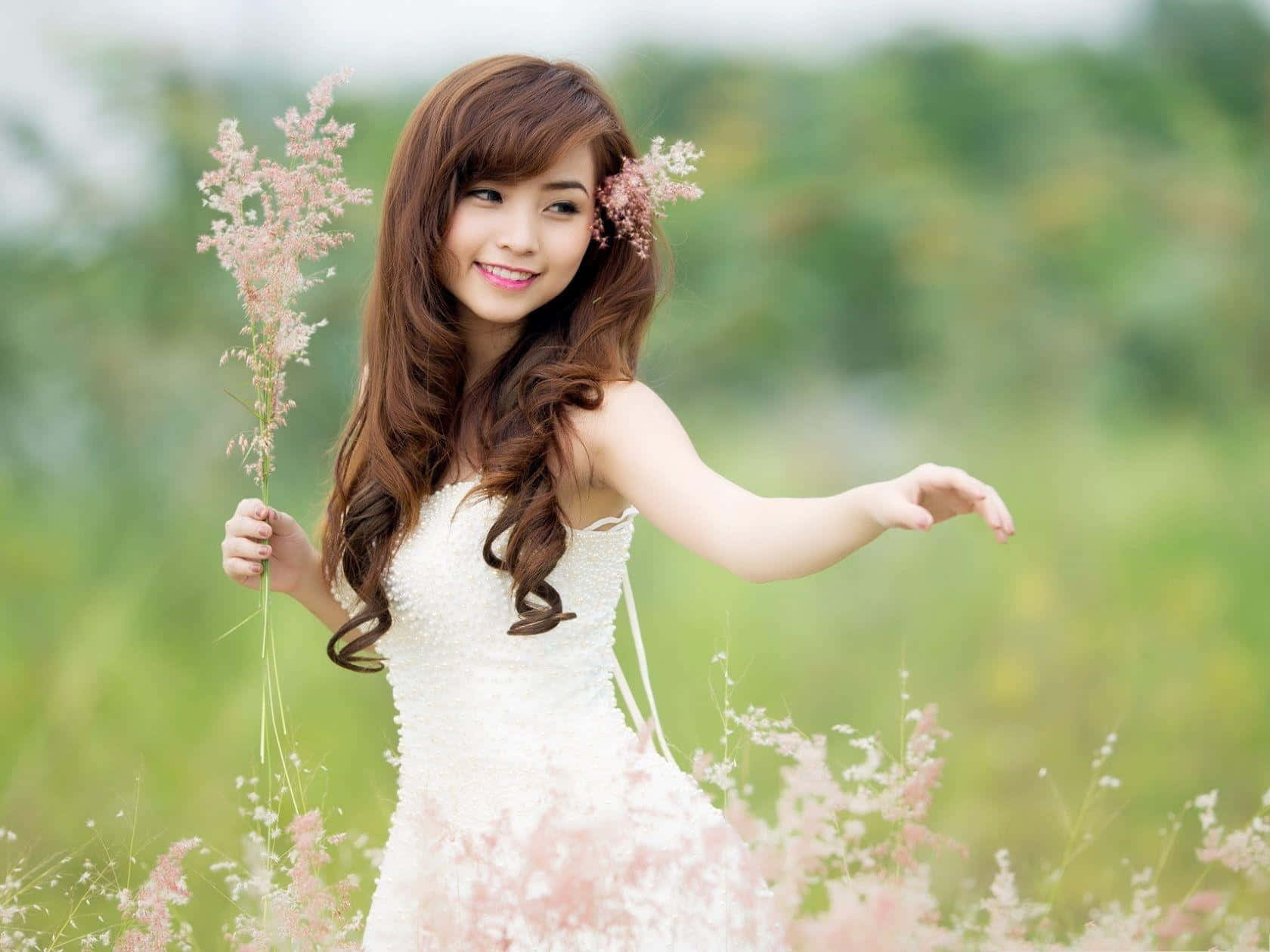 Beautiful Girls 桌布 壁紙 — 一位微笑的年輕女子身穿白色連衣裙，在陽光明媚的草地上手持粉紅色的草，背景是柔和的綠色散景。