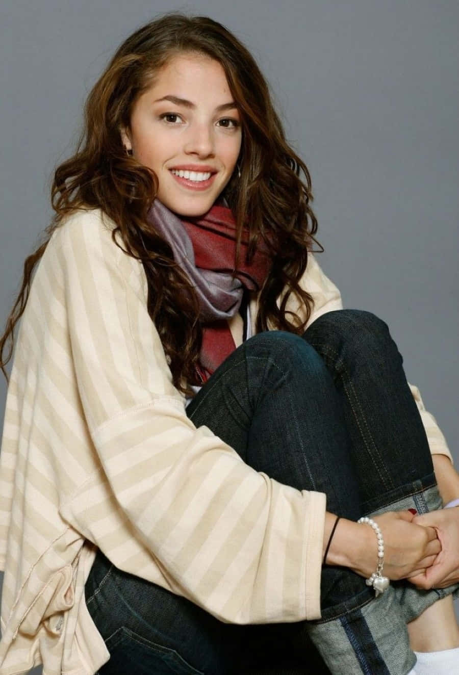 Olivia Thirlby 桌布 壁紙 — 一位微笑的年輕女性，波浪棕色頭髮，休閒地坐著，穿著條紋毛衣和圍巾，背景是純灰色。