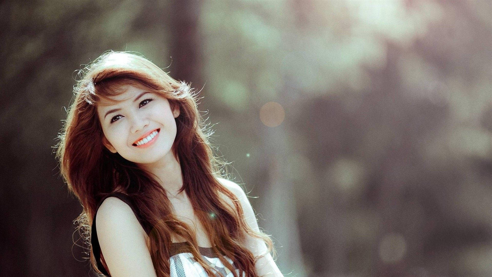 女孩 桌布 壁紙 — 一位微笑的年輕女性，長著紅棕色長髮，站在陽光普照的森林中，背景柔和散景。