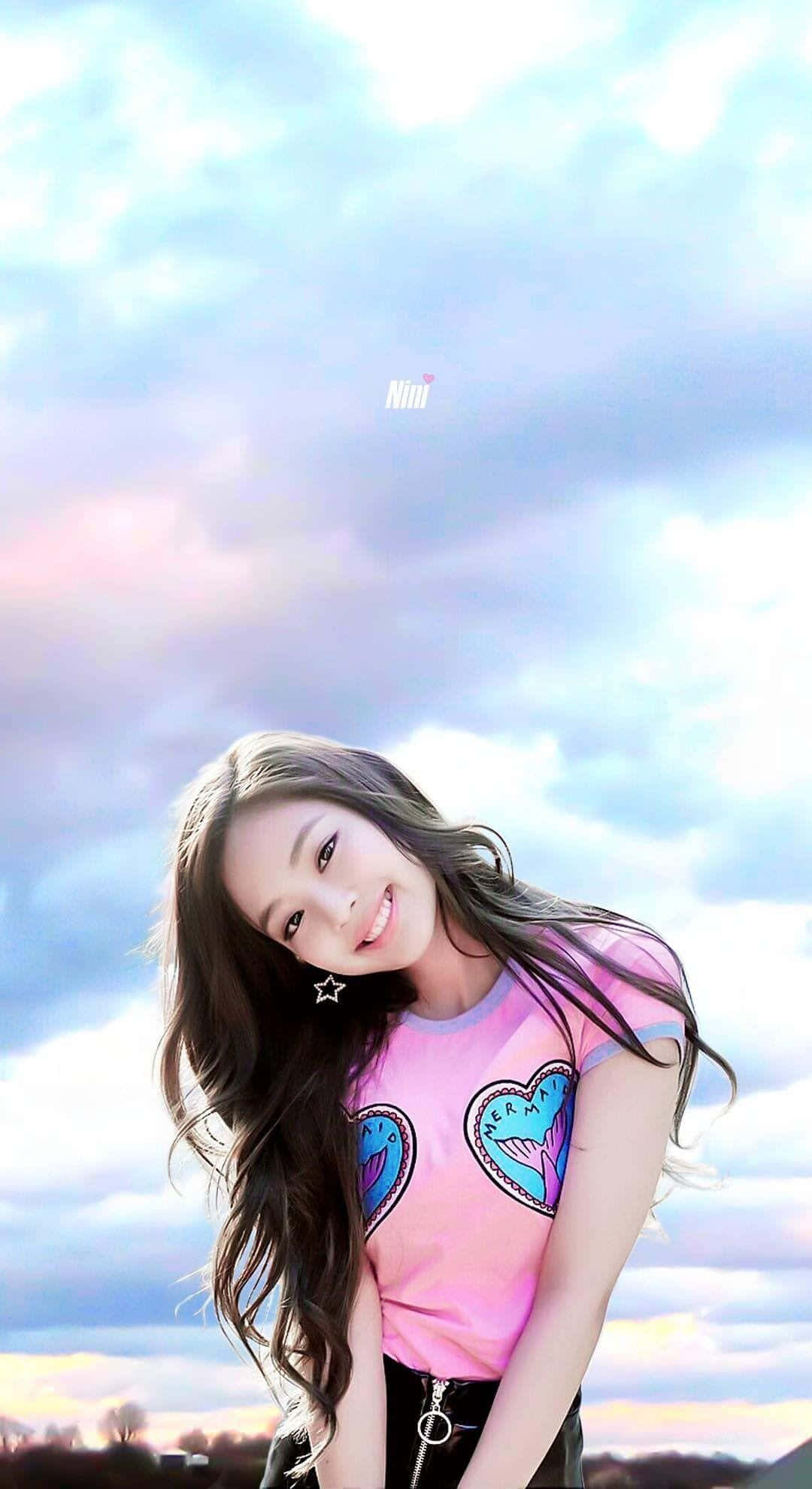 Jennie Kim 桌布 壁紙 — 一位微笑的年輕女性，長長的黑髮，穿著粉紅色心形印花T恤，背景是柔和粉彩的天空，有散落的雲朵。