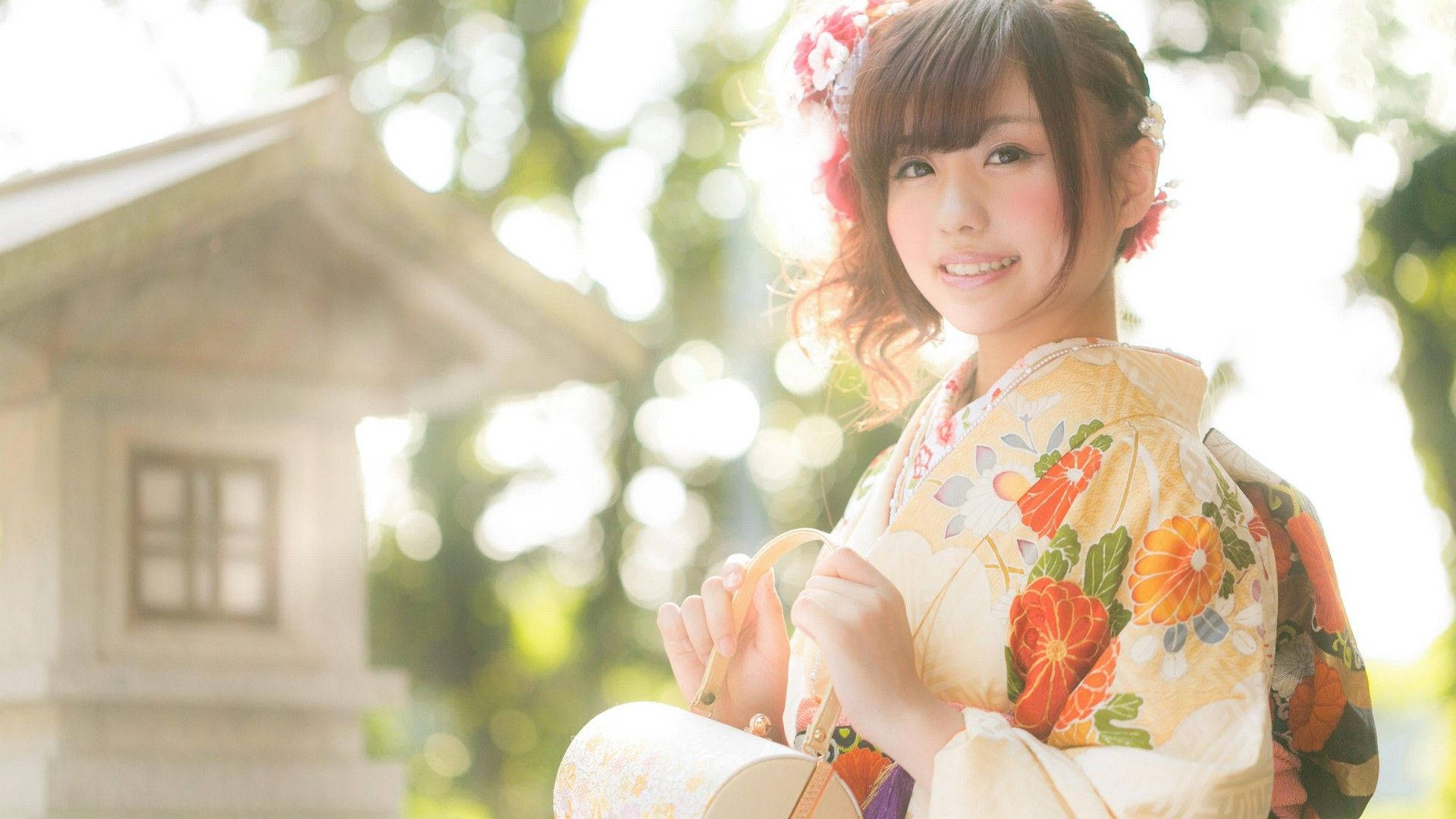 Beautiful Girl 桌布 壁紙 — 一位微笑的年輕女性穿著花卉和服，站在戶外石燈籠附近，柔和陽光穿過樹木灑落。