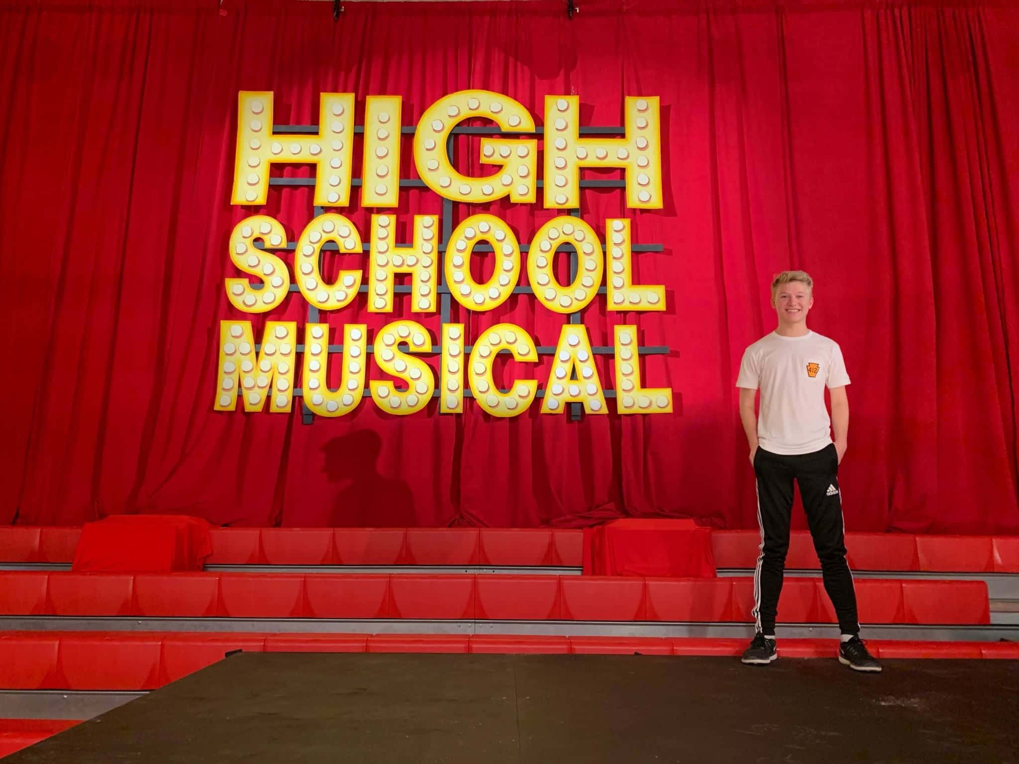 High School Musical 桌布 壁紙 — 一位微笑的年輕男子自信地站在紅色劇院舞台上，面前是發光的黃色「High School Musical」標誌。
