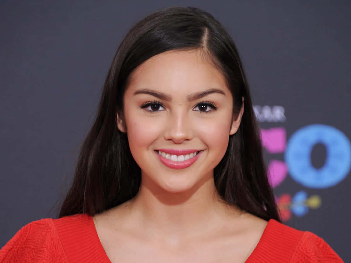 Olivia Rodrigo 桌布 壁紙 — 一位微笑的年輕黑髮女性，穿著紅色上衣，背景是色彩繽紛的活動場地。