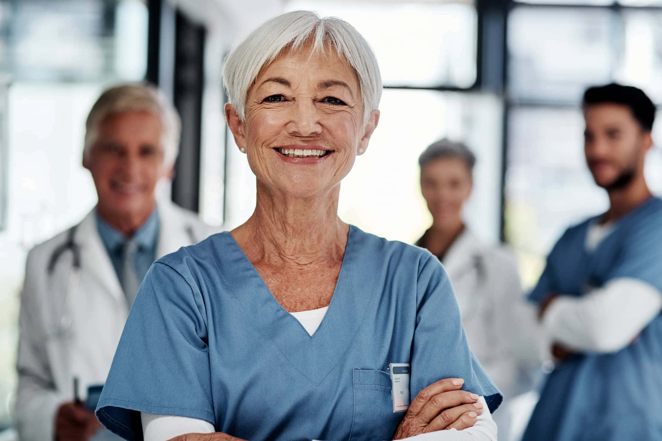 Nursing 桌布 壁紙 — 一位微笑的年長女護士穿著藍色工作服，自信地交叉雙臂站在明亮的醫院走廊，身後有模糊的同事。