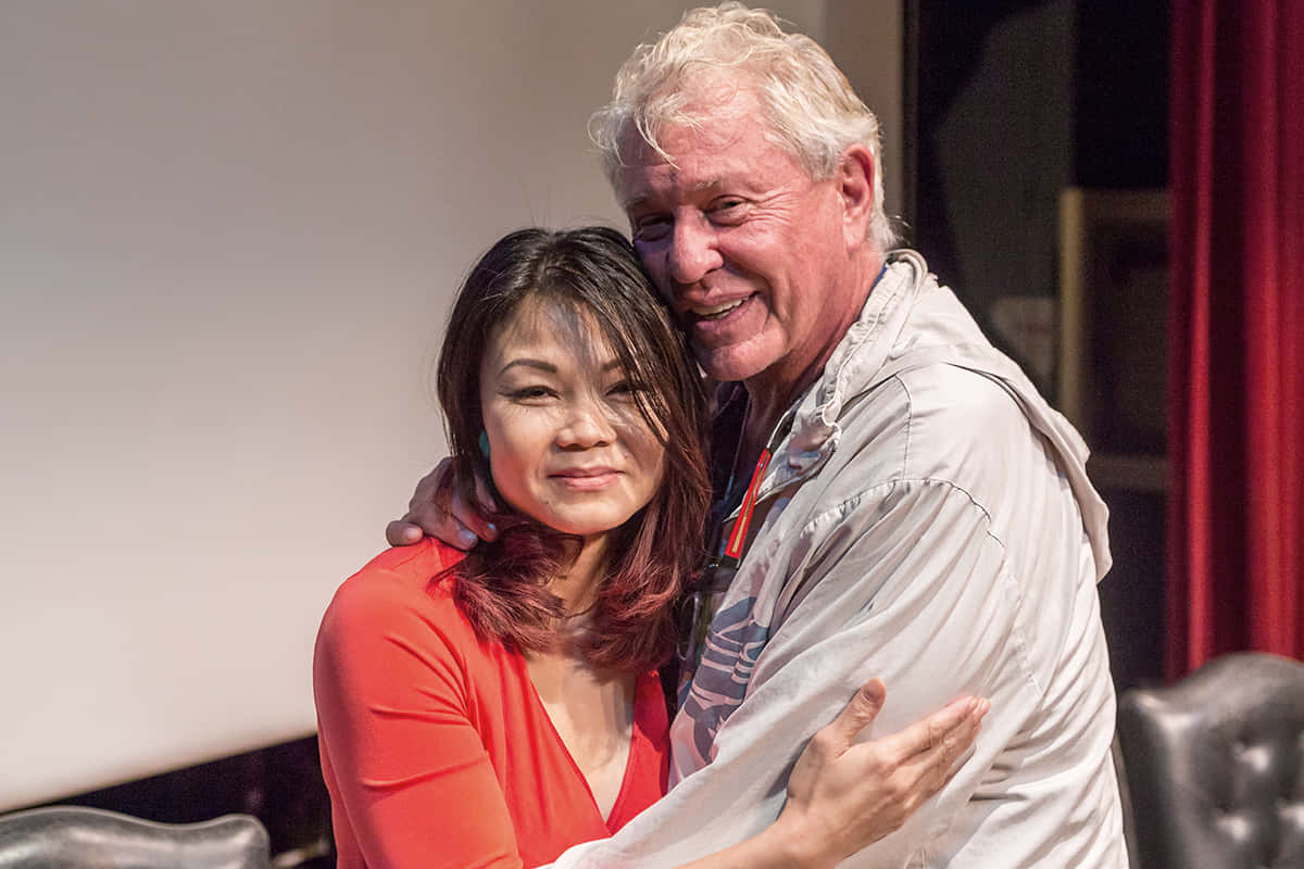 Tom Berenger 桌布 壁紙 — 一位微笑的年長男士擁抱著一位身穿紅色上衣的女性，背景是紅色窗簾和舞台燈光。