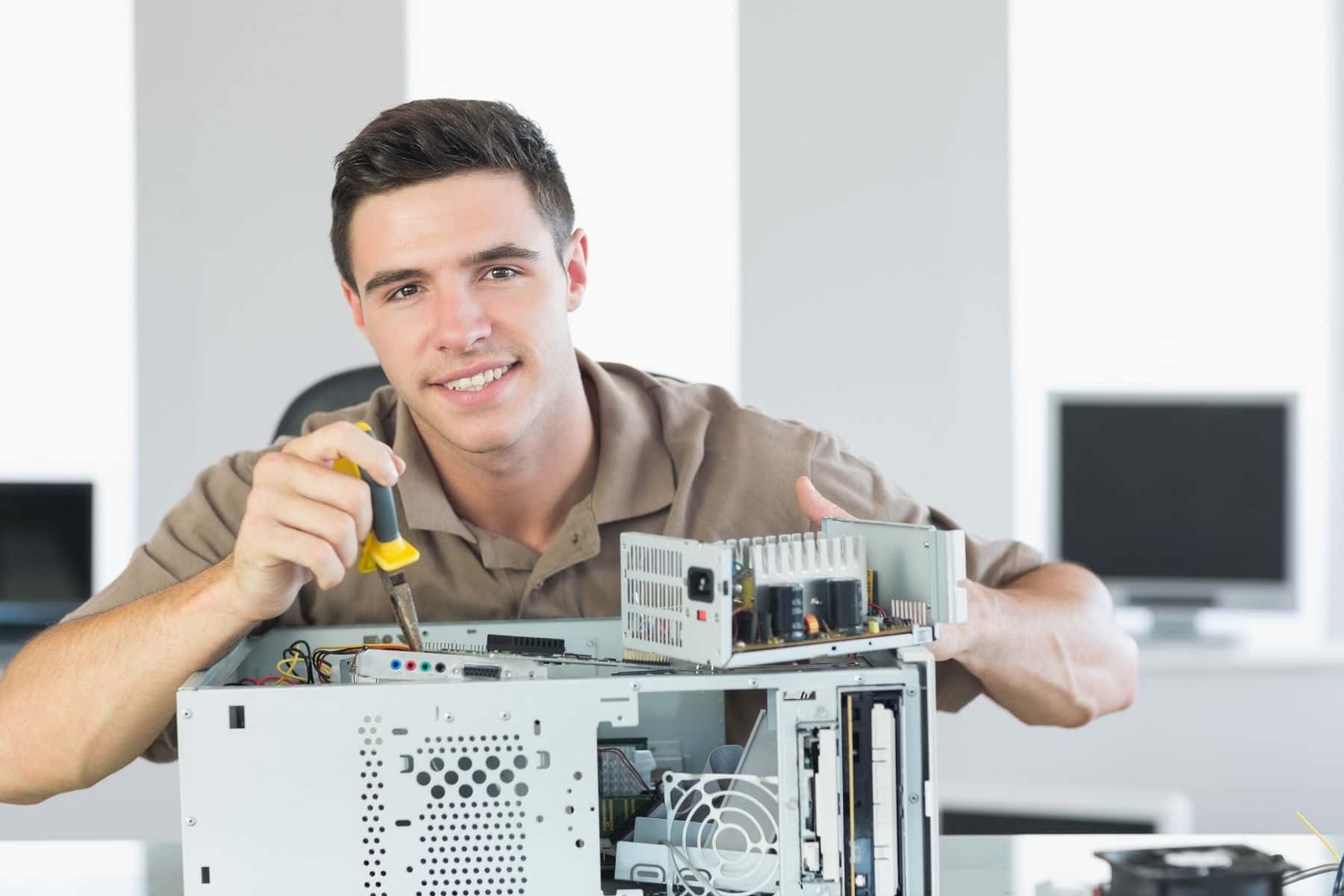 Engineer 桌布 壁紙 — 一位微笑的技術人員穿著棕色襯衫，在明亮的辦公室裡用螺絲刀修理打開的電腦機箱。