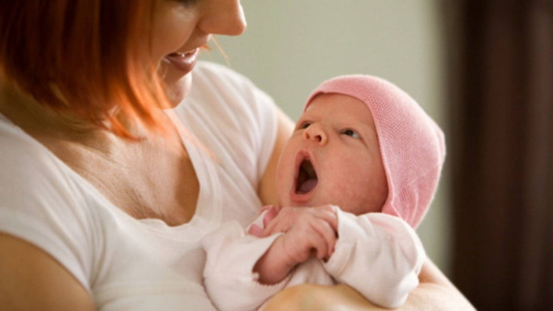 yawning 桌布 壁紙 — 一位微笑的母親在室內抱著她打呵欠的新生嬰兒，包裹在柔軟的粉紅色衣服中。