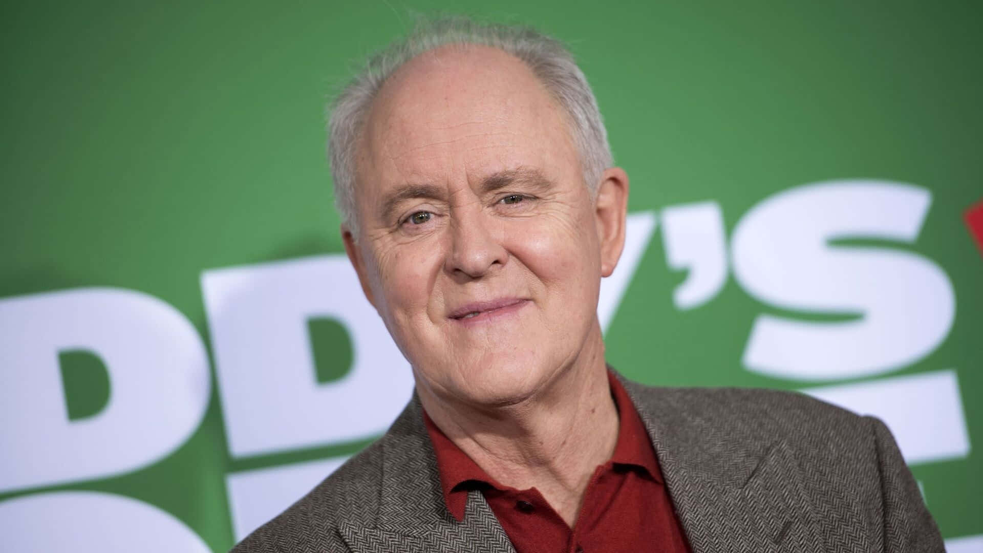 John Lithgow 桌布 壁紙 — 一位微笑的灰髮老年男子穿著紅色襯衫和粗花呢外套，在綠色背景前，帶有白色文字的close-up肖像。