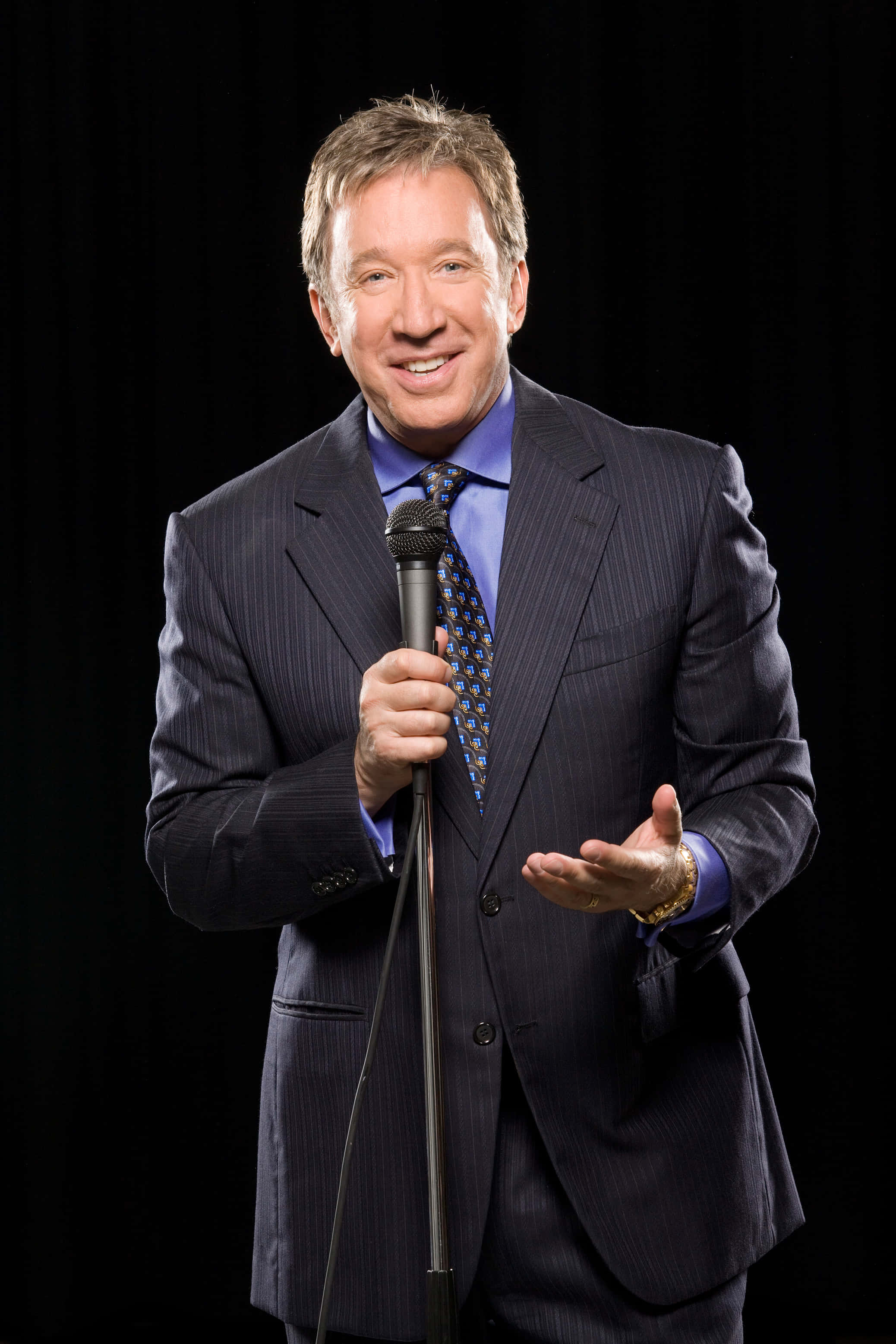 Tim Allen 桌布 壁紙 — 一位微笑的男士穿著條紋西裝，在黑色背景前握著麥克風，做出富有表現力的手勢。