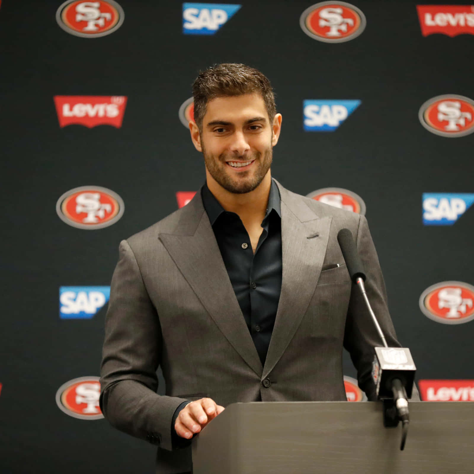 Jimmy Garoppolo 桌布 壁紙 — 一位微笑的男士穿著灰色西裝站在講台上，背景是舊金山49人和贊助商標誌。