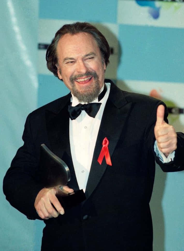 Rip Torn 桌布 壁紙 — 一位微笑的男士穿著禮服，佩戴紅色絲帶別針，豎起大拇指並手持獎項。