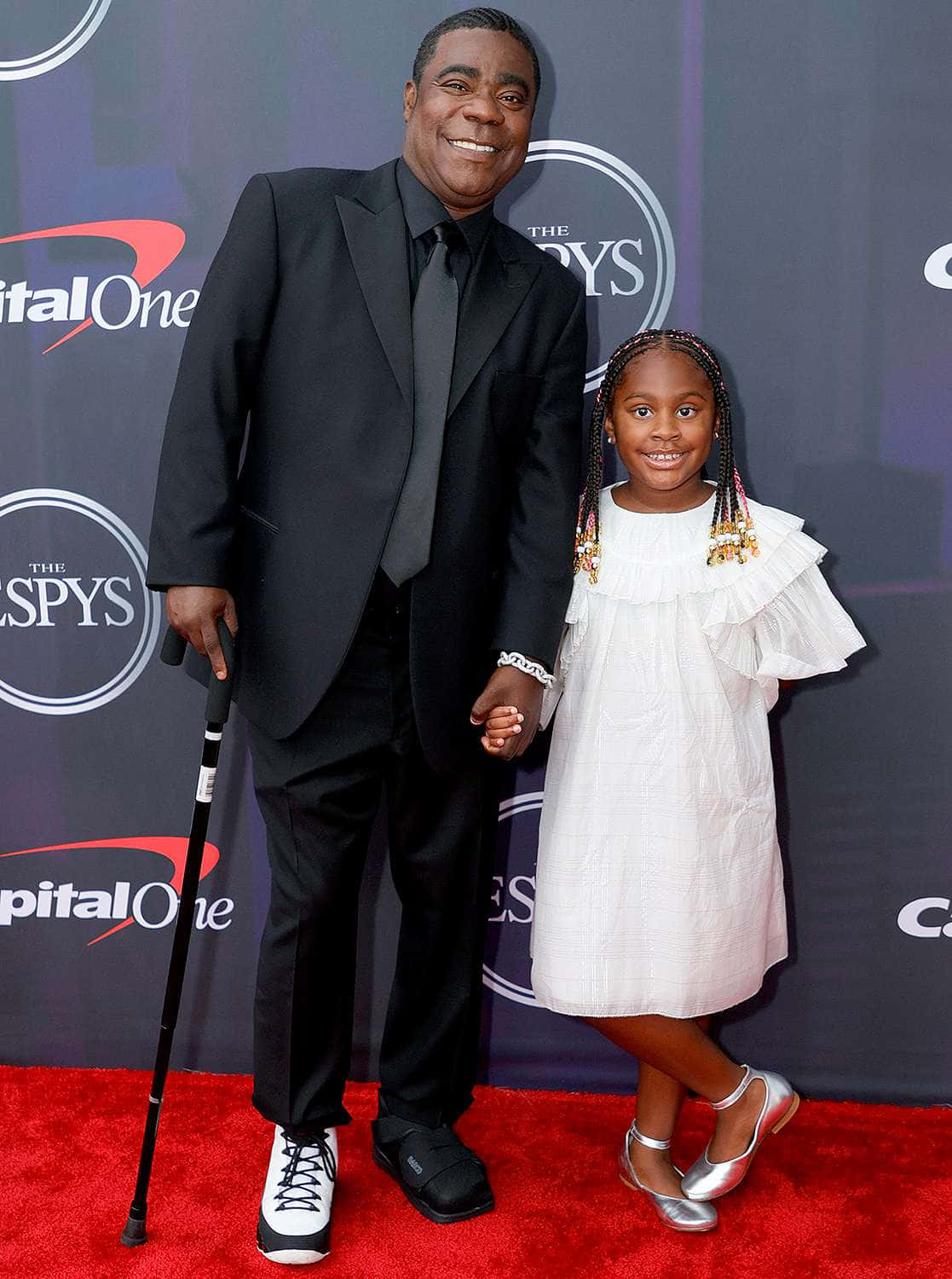 Tracy Morgan 桌布 壁紙 — 一位微笑的男士身穿黑色西裝、手持拐杖，在頒獎典禮的紅地毯上與一位身穿白色連衣裙的小女孩手牽手。