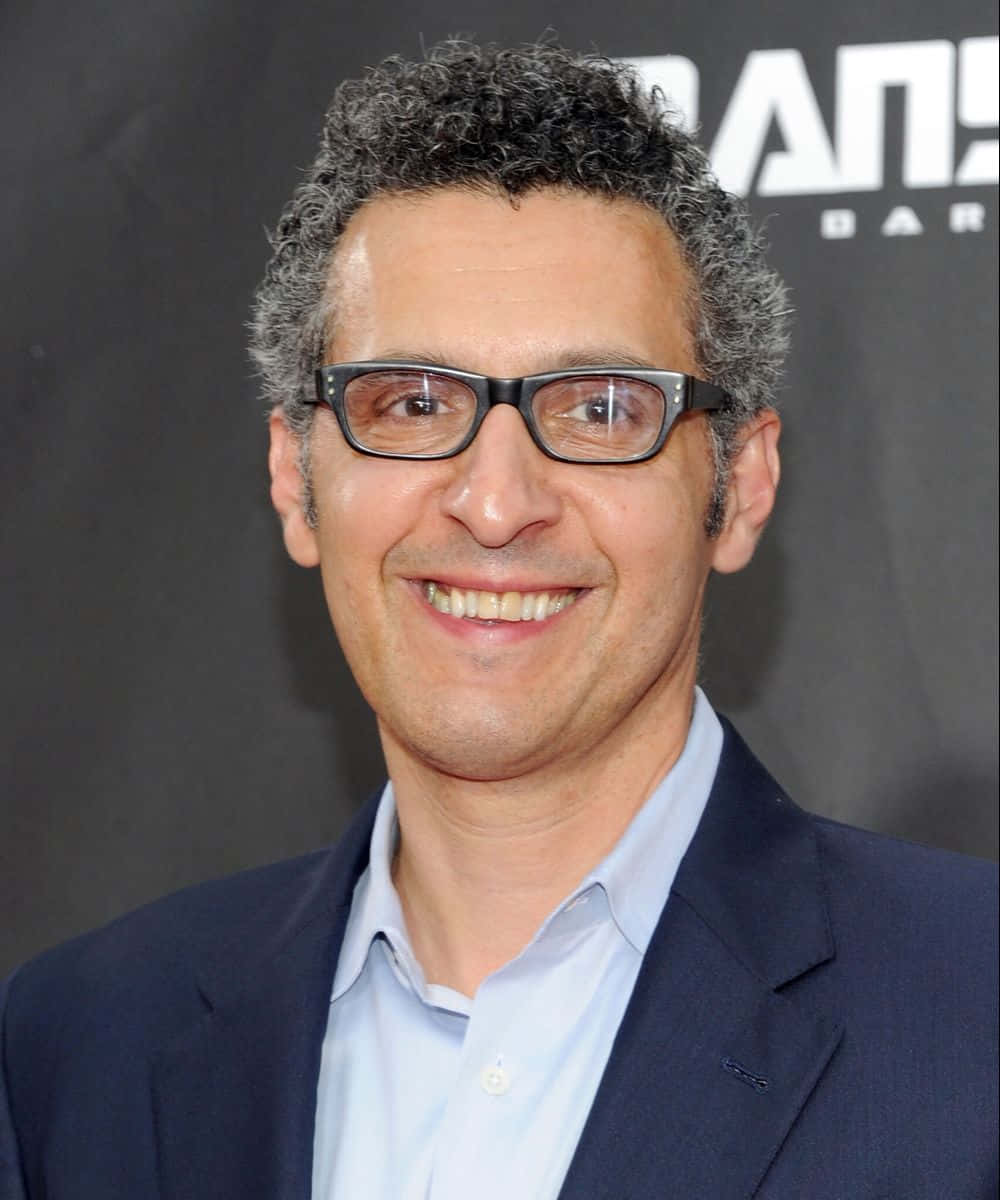 John Turturro 桌布 壁紙 — 一位微笑的男子，灰色捲髮，戴黑色眼鏡，身穿海軍藍西裝和淺藍色襯衫，背景為深色。