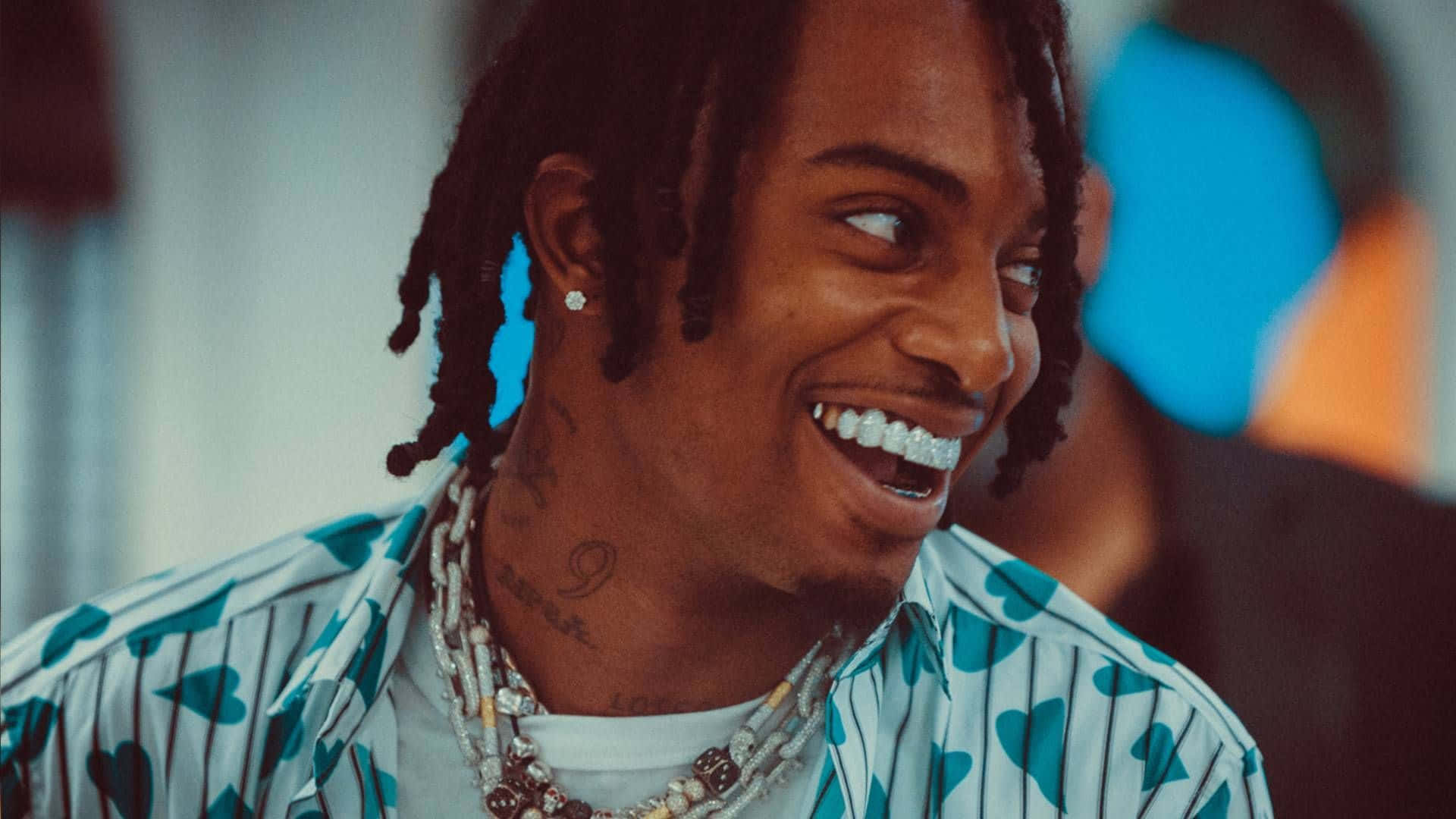 playboi carti 桌布 壁紙 — 一位微笑的男子，留著辮子頭和紋身，穿著心形圖案襯衫和層疊項鍊，背景是色彩繽紛的模糊效果。
