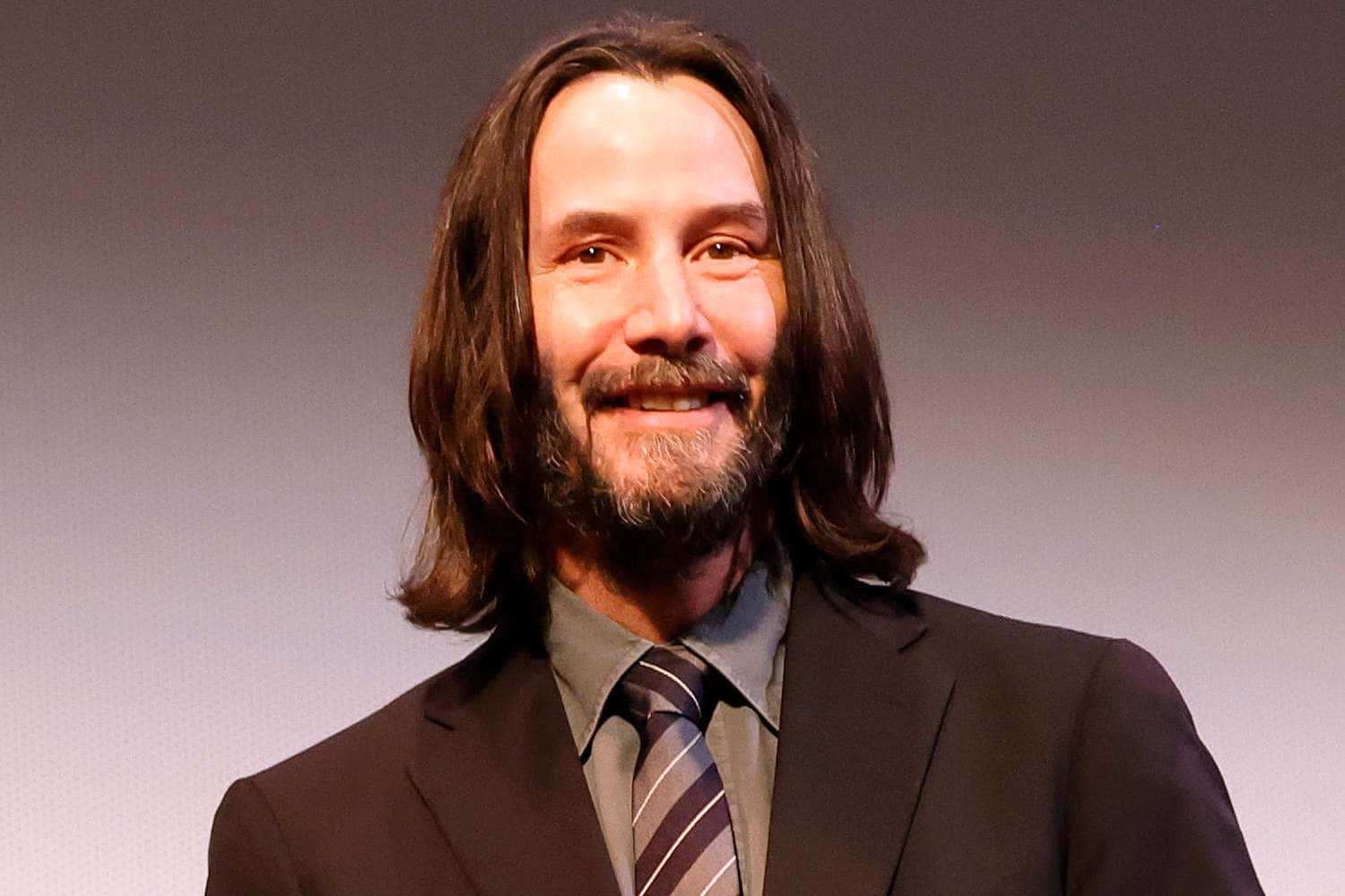 Keanu Reeves 桌布 壁紙 — 一位微笑的男子，留著長長的棕色頭髮和鬍鬚，穿著深色西裝和條紋領帶，背景是純灰色。