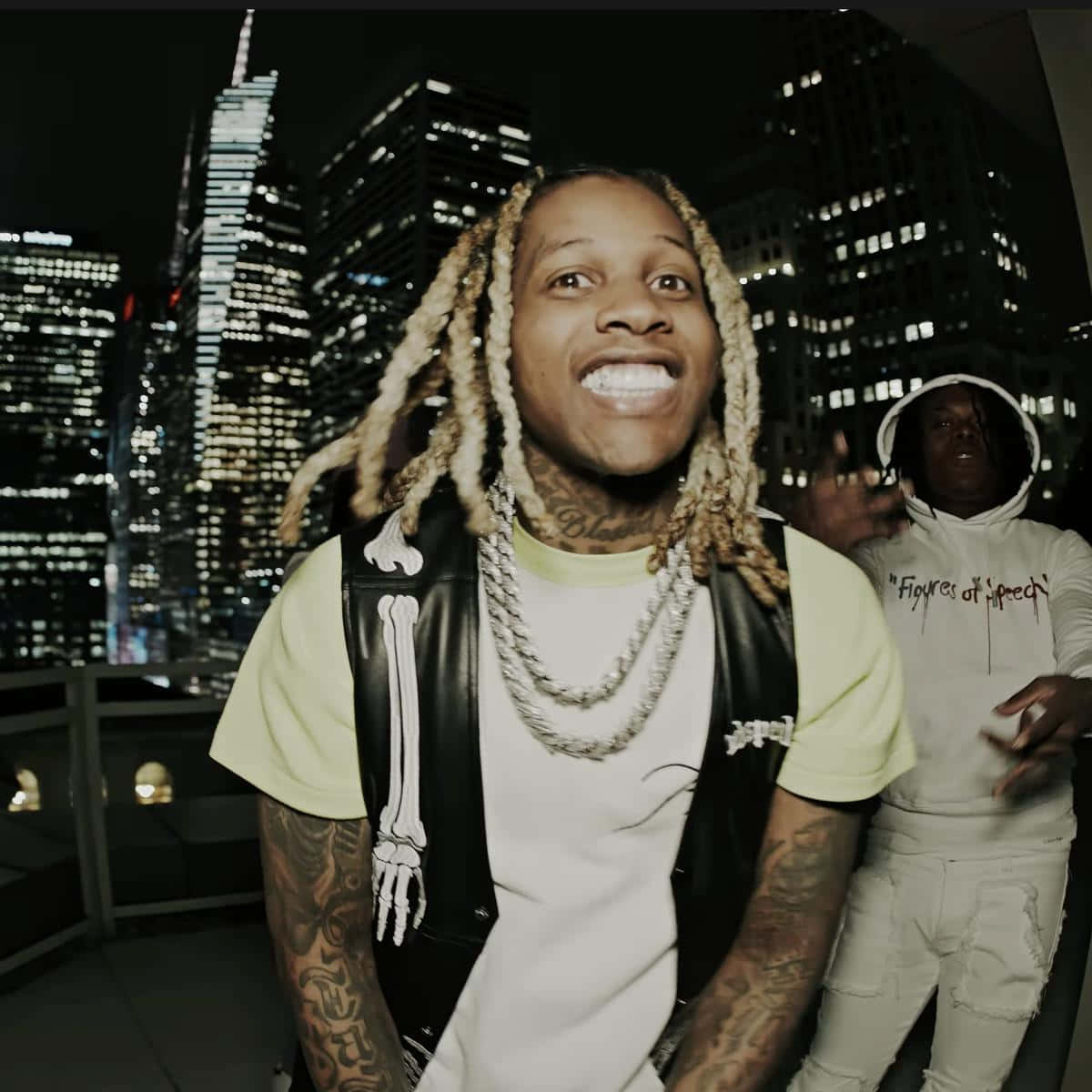 lil durk 桌布 壁紙 — 一位微笑的男子，編著辮子頭髮並有紋身，在夜晚的屋頂上以燈火通明的城市天際線為背景擺姿勢。