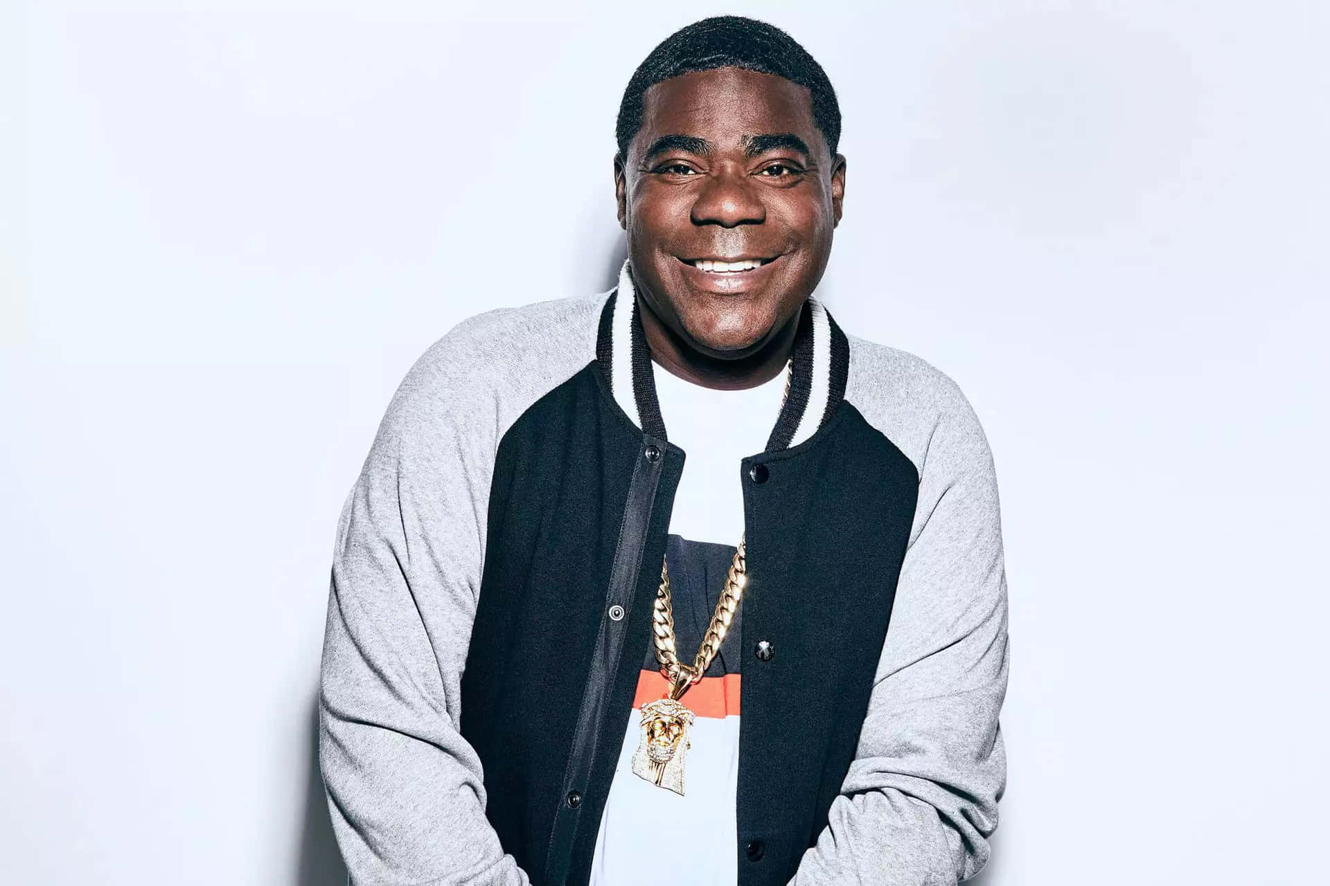 Tracy Morgan 桌布 壁紙 — 一位微笑的男子，身穿黑灰色校隊夾克，戴著金鏈項鍊，背景為純白色。