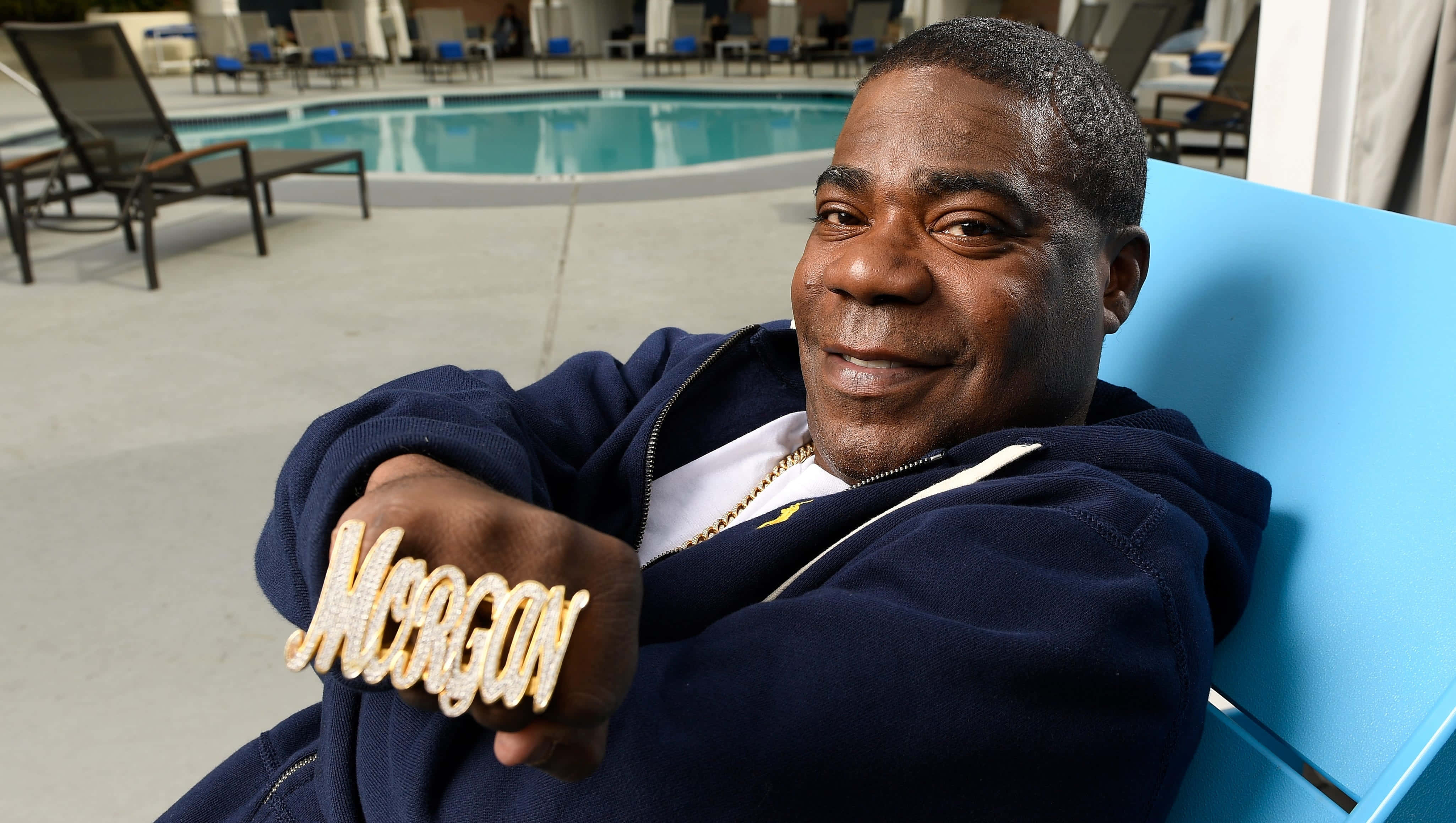Tracy Morgan 桌布 壁紙 — 一位微笑的男子在泳池旁的藍色椅子上休息，穿著海軍藍連帽衫和一枚鑲有鑽石的 'MORGAN' 大戒指。