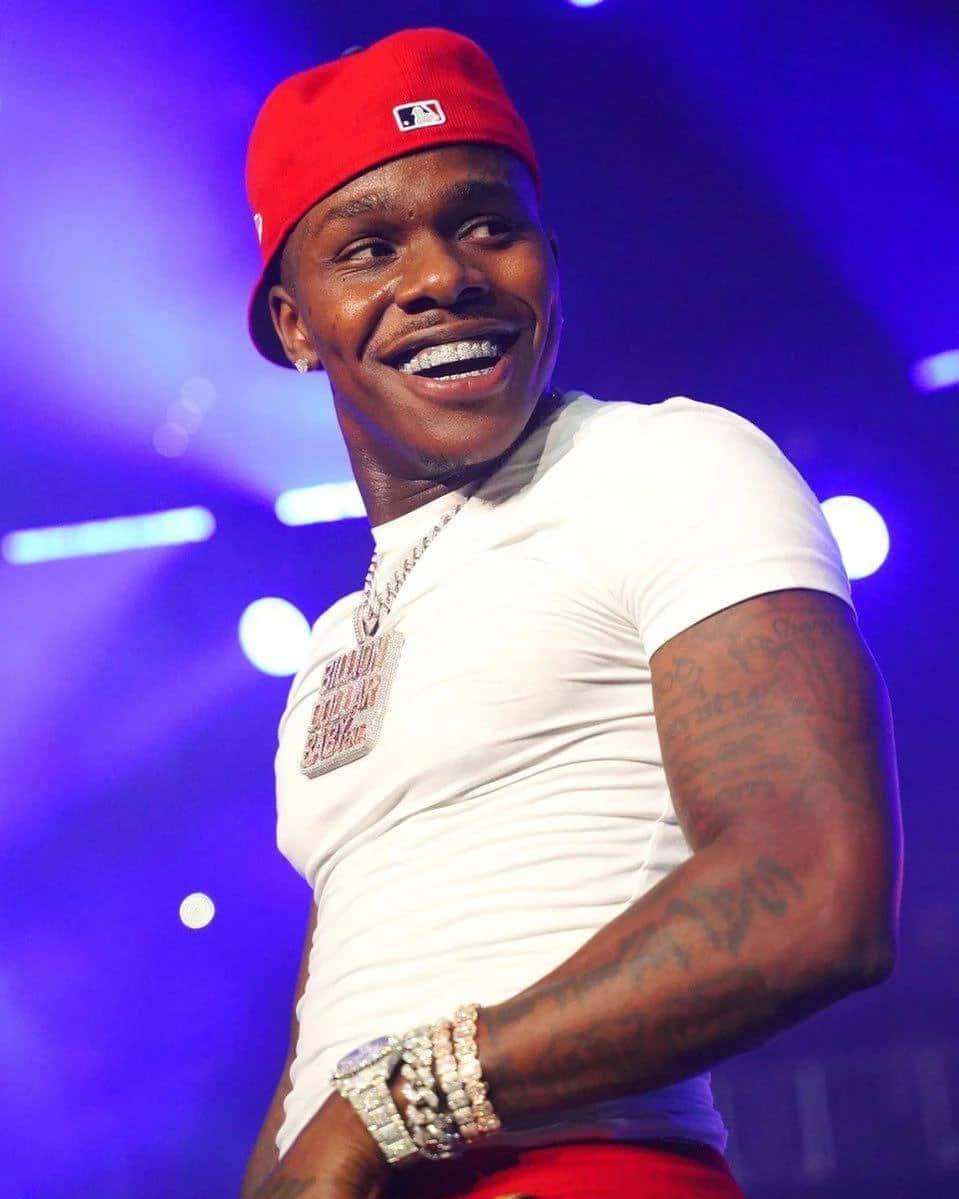 Dababy 桌布 壁紙 — 一位微笑的男子戴著紅色帽子、穿著白色襯衫，在充滿活力的藍色舞台燈光下表演，身上可見紋身和珠寶。