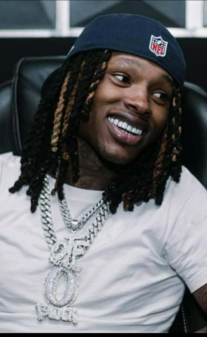 King Von 桌布 壁紙 — 一位微笑的男子梳著辮子，戴著海軍藍NFL帽子和閃亮的'OF O'鏈條，坐在黑色椅子上。