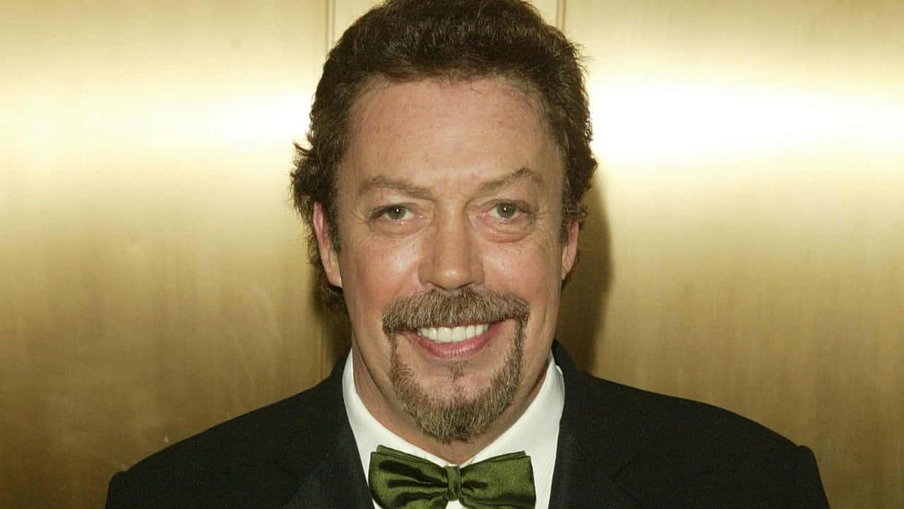 Tim Curry 桌布 壁紙 — 一位微笑的男子留著鬍鬚和山羊鬍，身穿黑色西裝和綠色領結，背景是金色牆壁。