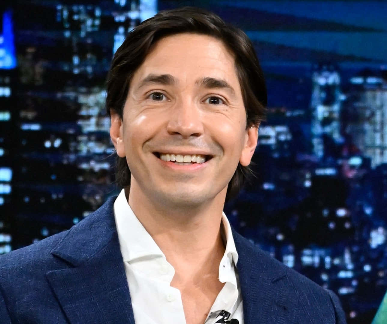 Justin Long 桌布 壁紙 — 一位微笑的男子穿著藍色西裝外套和白色襯衫，站在模糊的夜間城市景觀背景前。