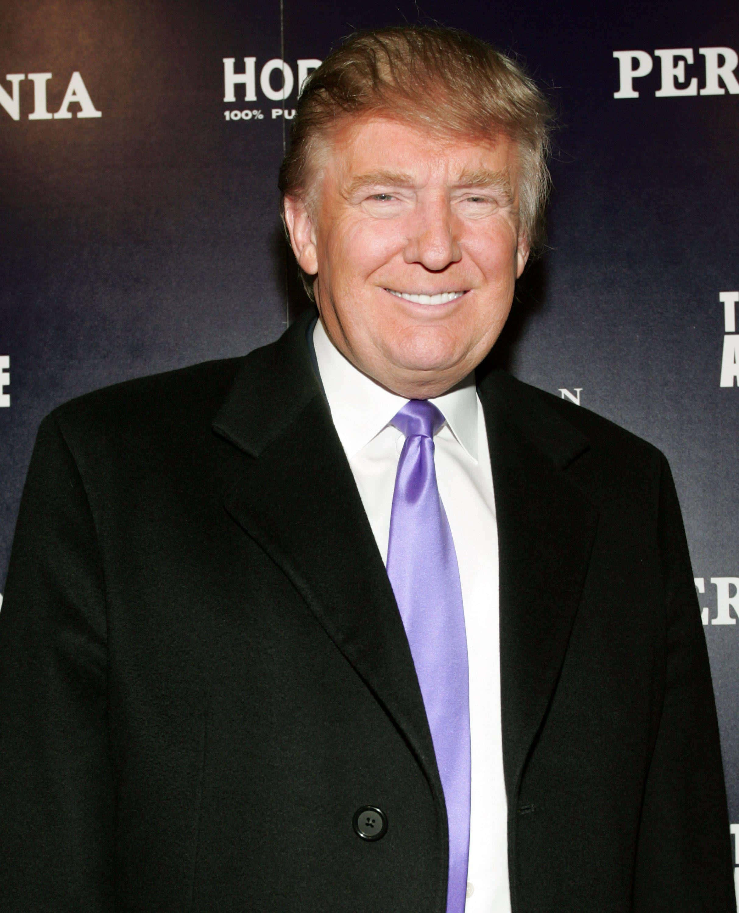trump 桌布 壁紙 — 一位微笑的男子穿著黑色外套和紫色領帶，在活動中於深色品牌背景前擺姿勢。
