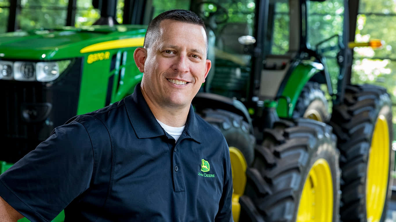 John Deere 桌布 壁紙 — 一位微笑的男子身穿John Deere Polo衫，自信地站在一輛帶有大型黑色輪胎的綠色和黃色拖拉機前。