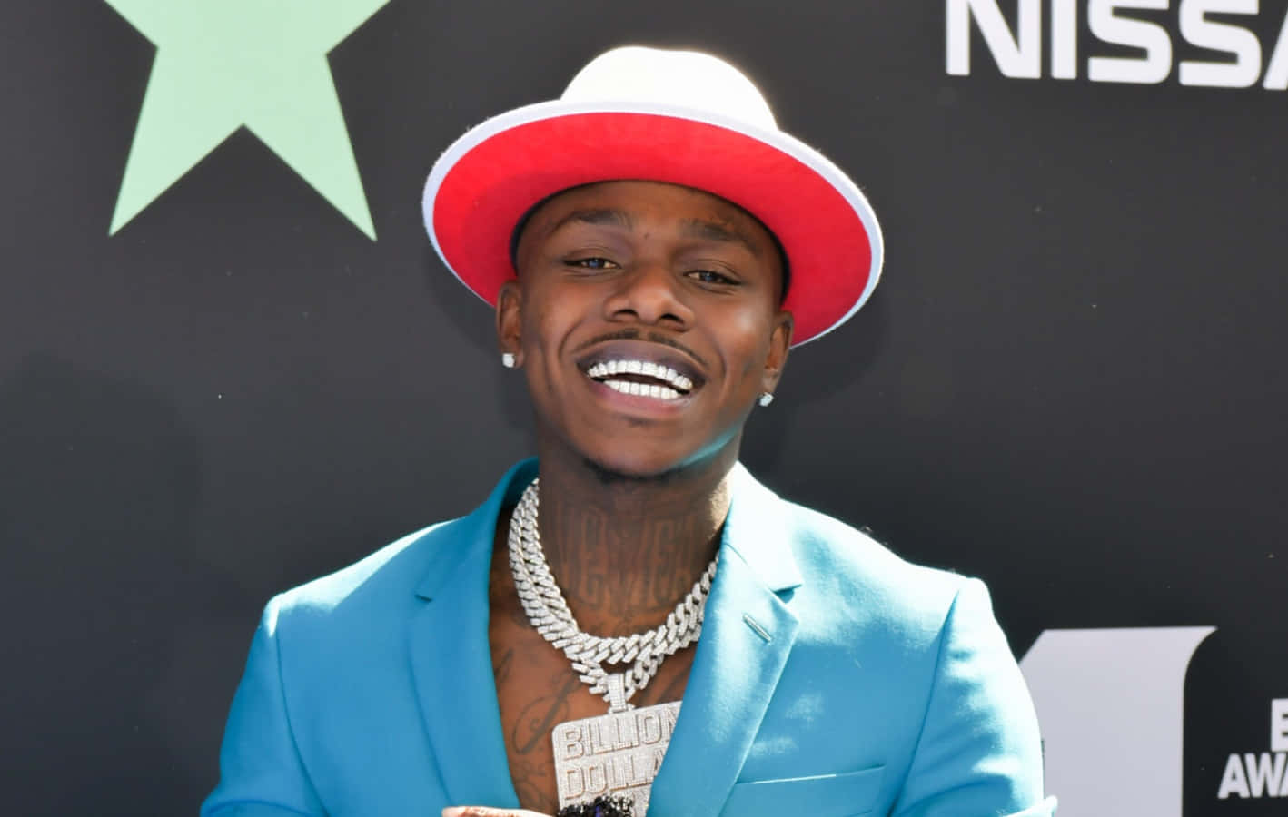 Dababy 桌布 壁紙 — 一位微笑的男子身穿亮藍色西裝和紅色帽子，在日產標誌背景的頒獎活動中自信地擺姿勢。