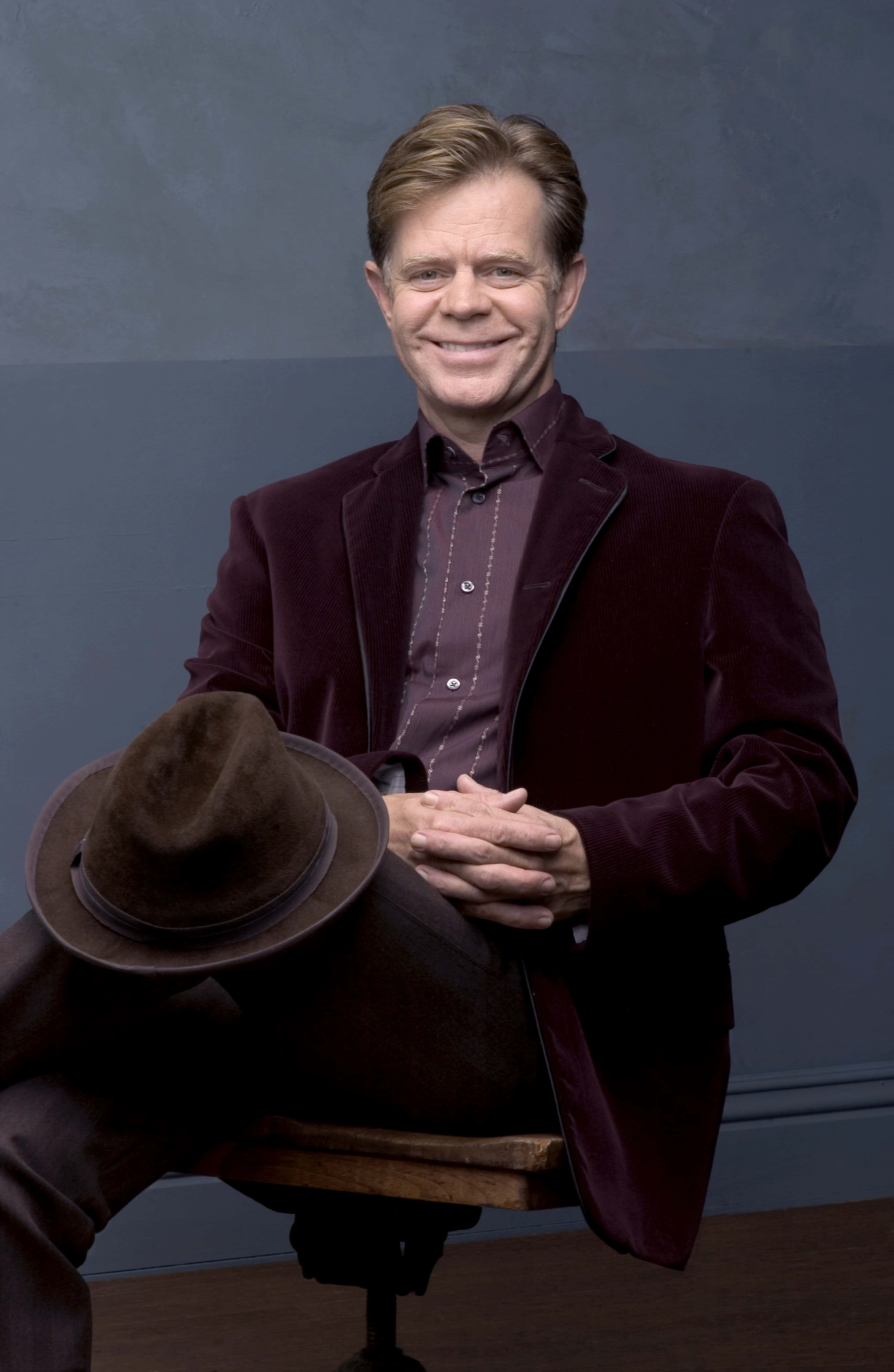 william h. macy 桌布 壁紙 — 一位微笑的男子身穿栗紅色天鵝絨西裝，坐在凳子上，手持一頂棕色軟呢帽，背景為柔和的灰色。