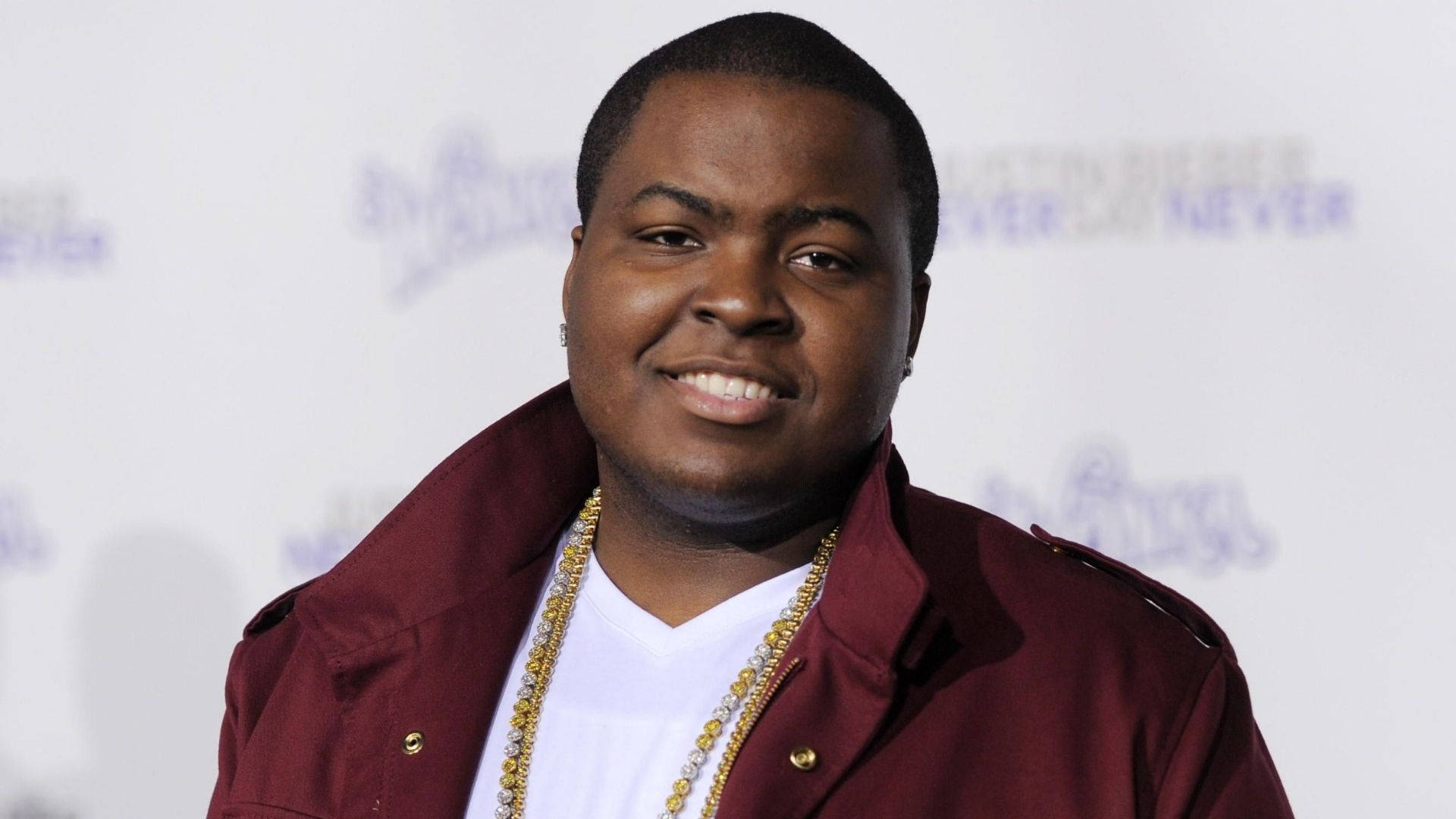 Sean Kingston Sweet Smile 壁紙 — 一位微笑的男子身穿栗色外套和金色鏈條，在帶有模糊標誌的白色活動背景前擺姿勢。