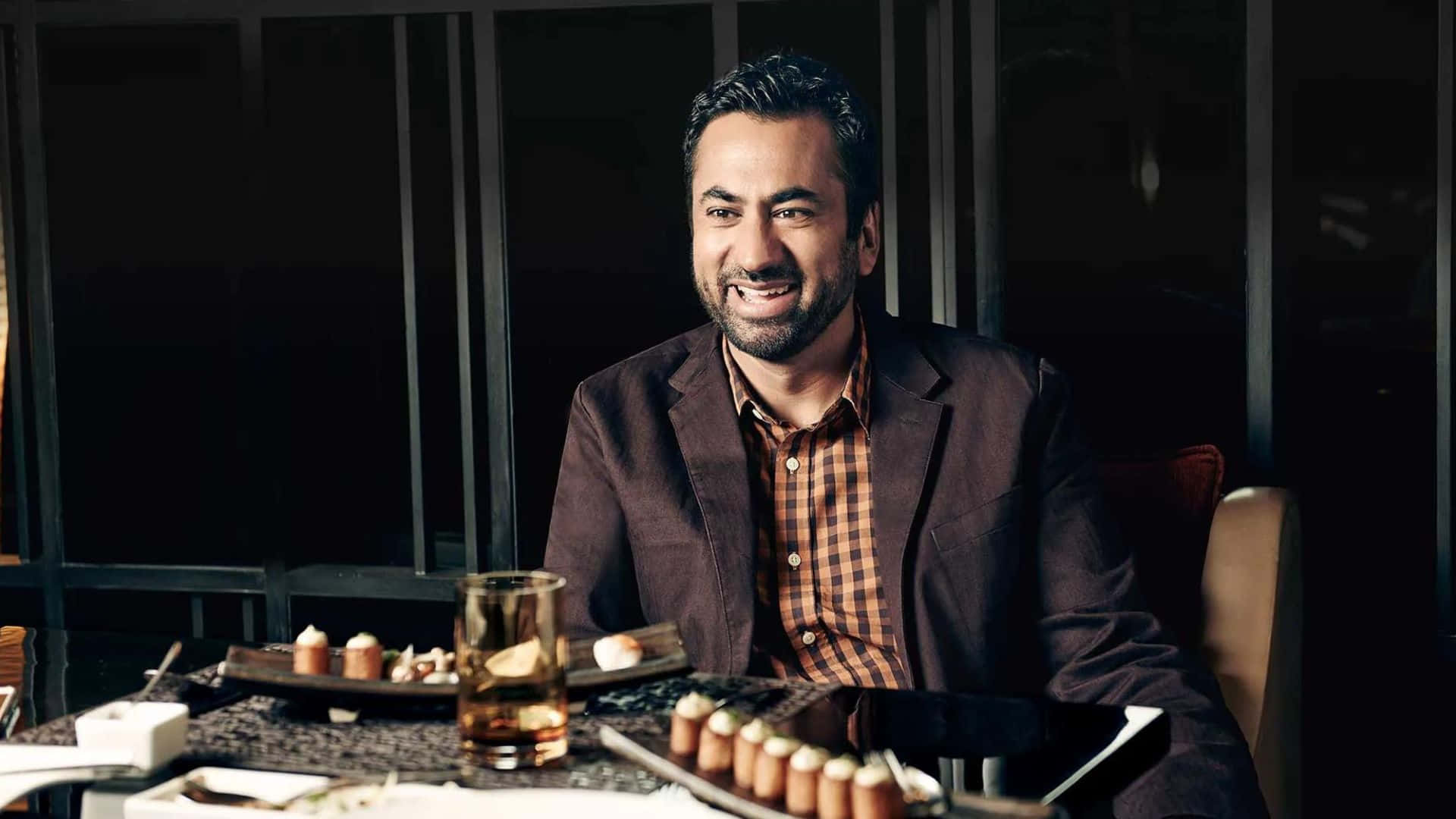 Kal Penn 桌布 壁紙 — 一位微笑的男子身穿棕色西裝外套，坐在燈光昏暗的餐桌前，面前擺放著開胃菜和一杯飲料。