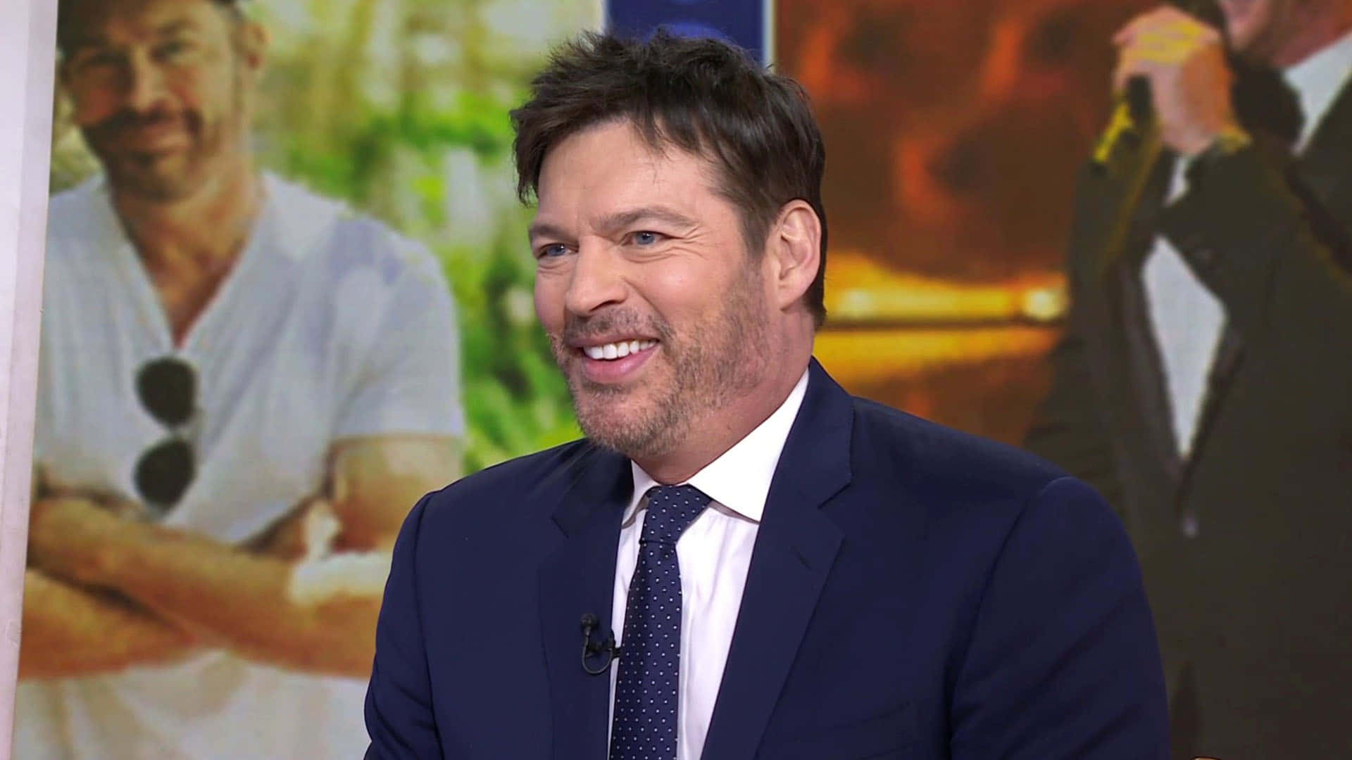 Harry Connick Jr 桌布 壁紙 — 一位微笑的男子身穿海軍藍西裝配波點領帶，在模糊的宣傳背景佈景中發言。