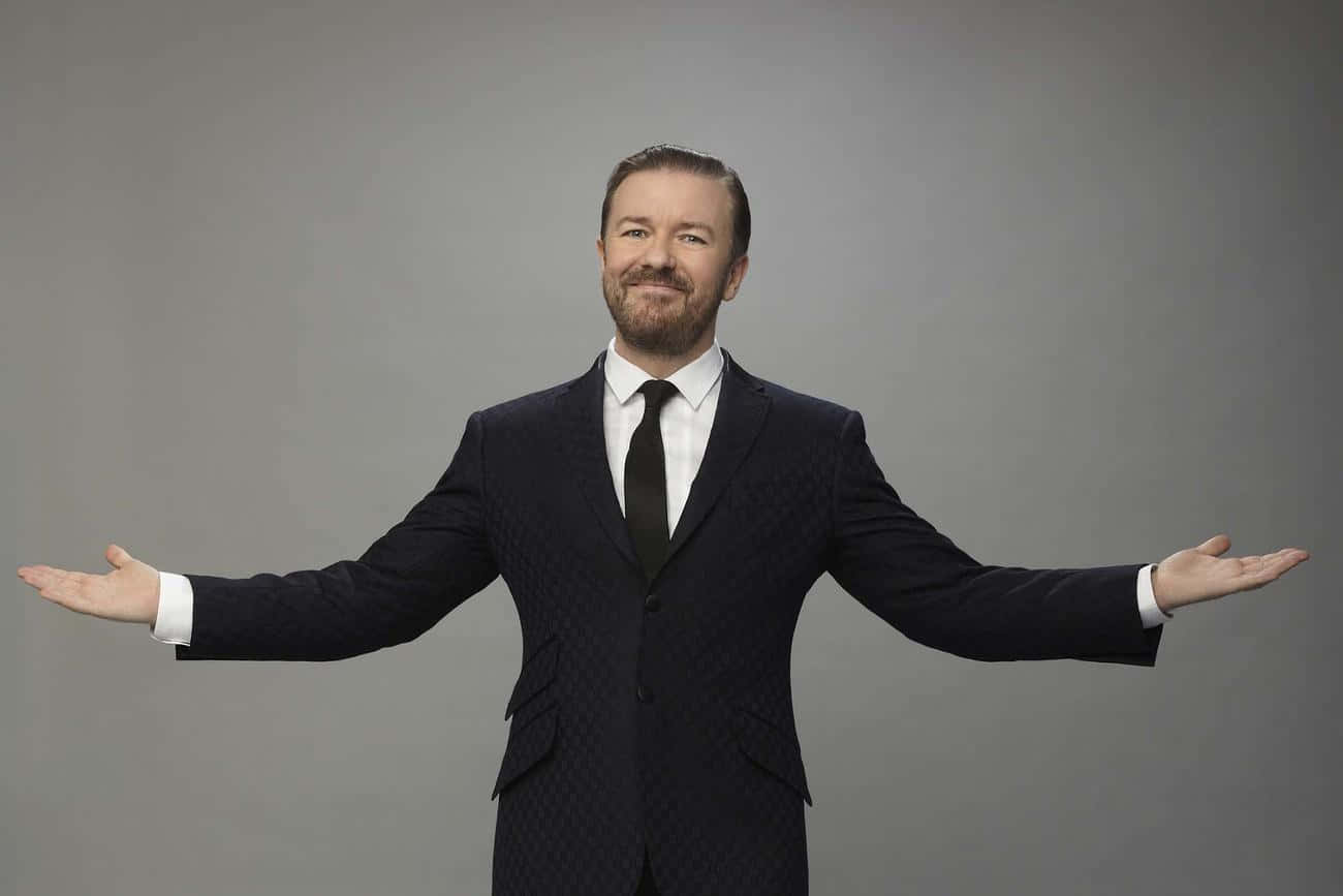 ricky gervais 桌布 壁紙 — 一位微笑的男子身穿深色圖案西裝，雙臂張開，站在純灰色攝影棚背景前。