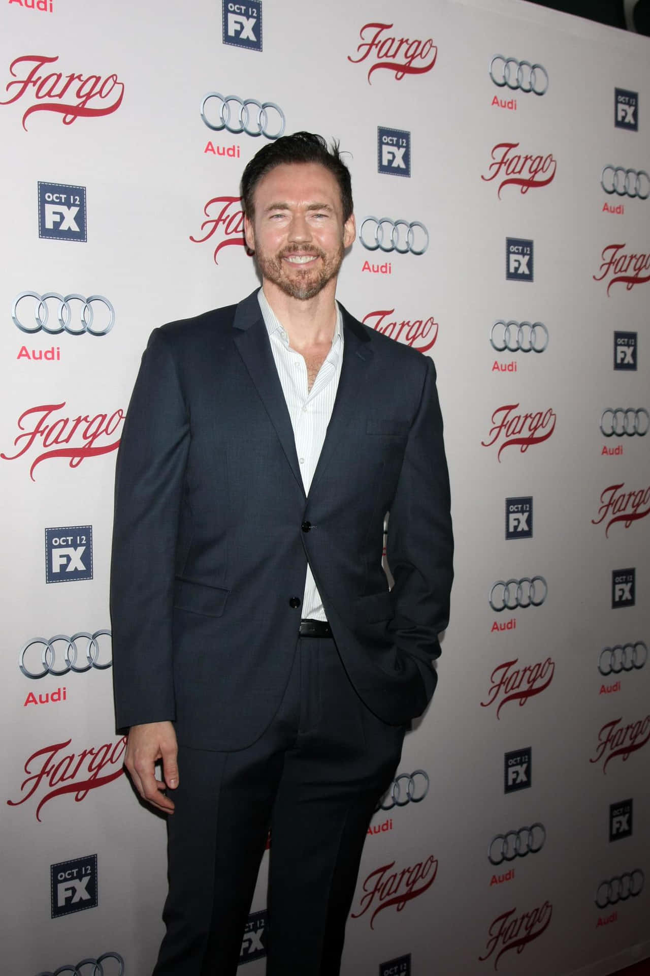 Kevin Durand 桌布 壁紙 — 一位微笑的男子身穿深色西裝，自信地站在紅地毯活動上，身後有 'Fargo' 和 'Audi' 標誌。