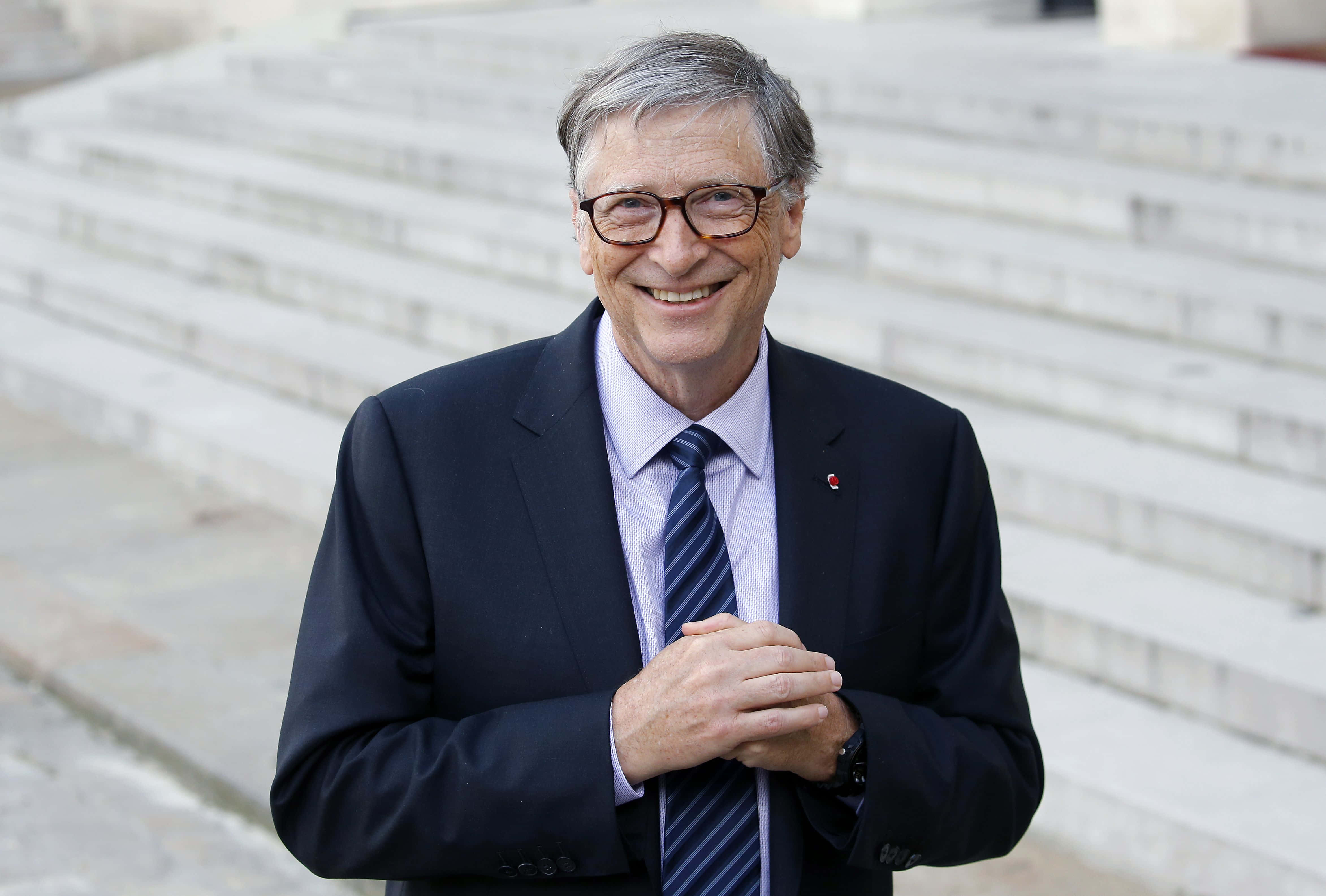 Bill Gates 桌布 壁紙 — 一位微笑的男子身穿深色西裝和眼鏡，在明亮的日光下自信地站在寬闊的石階前。