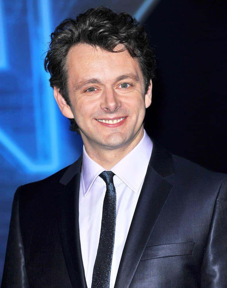 Michael Sheen 桌布 壁紙 — 一位微笑的男子身穿深色西裝和領帶，在活動中站在發光的藍色背景前。