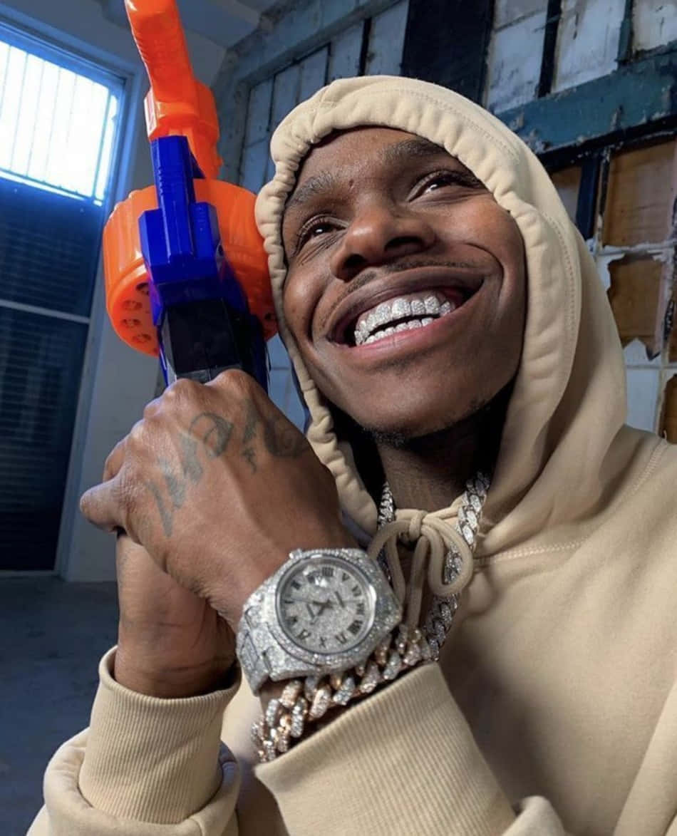 Dababy 桌布 壁紙 — 一位微笑的男子身穿米色連帽衫，手持橙色和藍色的水槍，佩戴鑲鑽手錶和鏈條。
