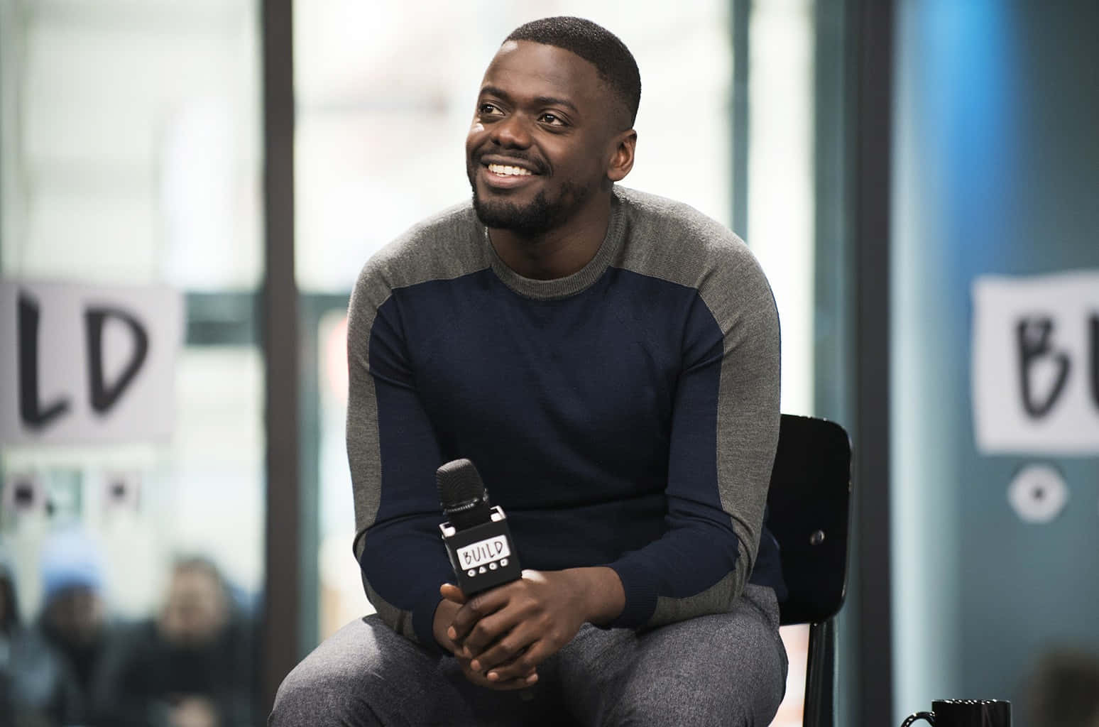Daniel Kaluuya 桌布 壁紙 — 一位微笑的男子身穿藍灰色毛衣，坐在現代工作室環境中，手持麥克風。