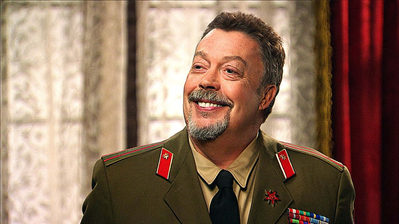 Tim Curry 桌布 壁紙 — 一位微笑的男子身穿裝飾性的橄欖綠軍裝，配有紅色肩章和勳章，站在圖案窗簾和紅色帷幔前。