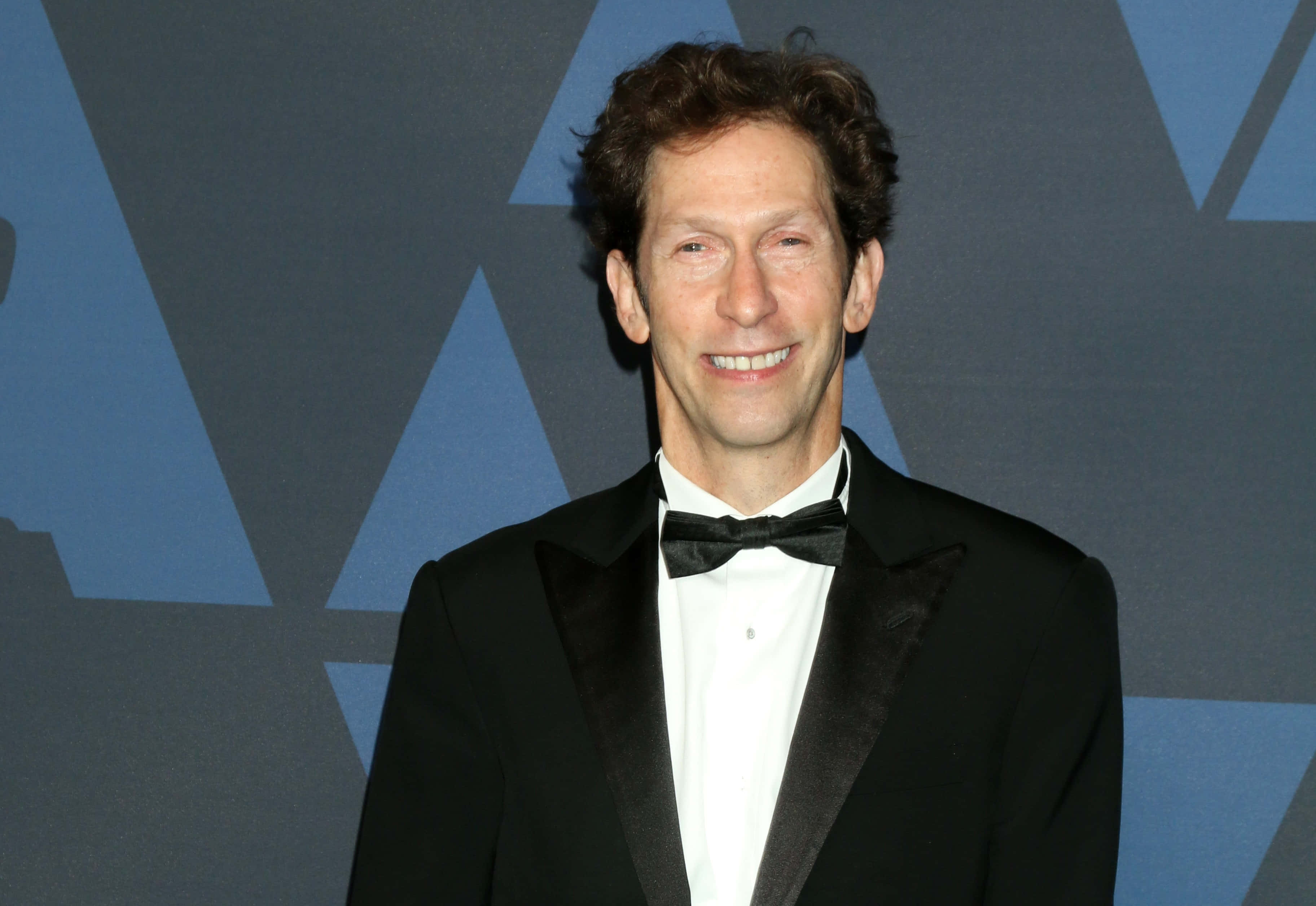 Tim Blake Nelson 桌布 壁紙 — 一位微笑的男子身穿黑色燕尾服配蝴蝶結，站在藍色幾何圖案背景前。