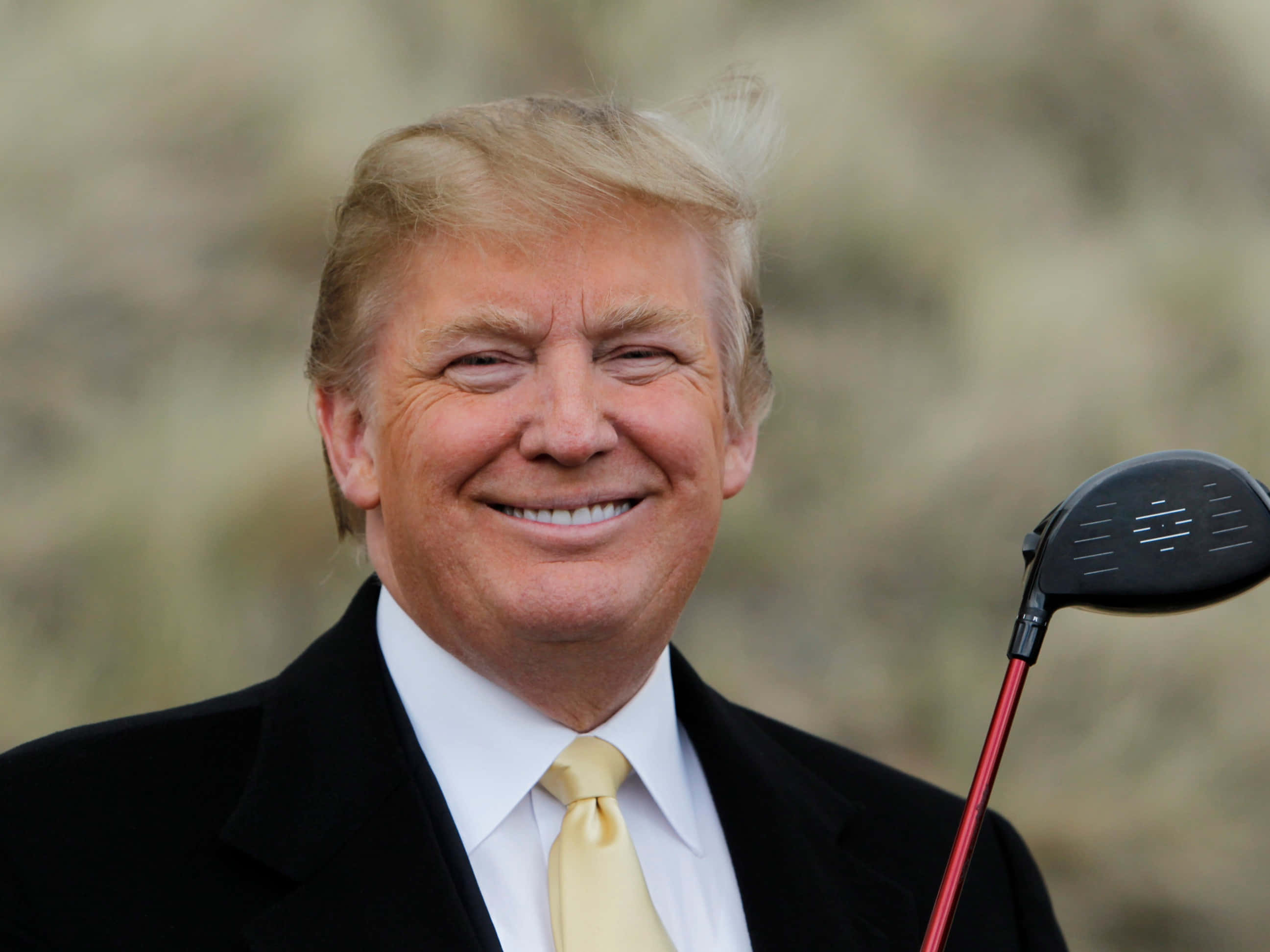 trump 桌布 壁紙 — 一位微笑的男子身穿黑色西裝、繫著黃色領帶，在模糊的自然背景前握著一支紅色高爾夫球桿。