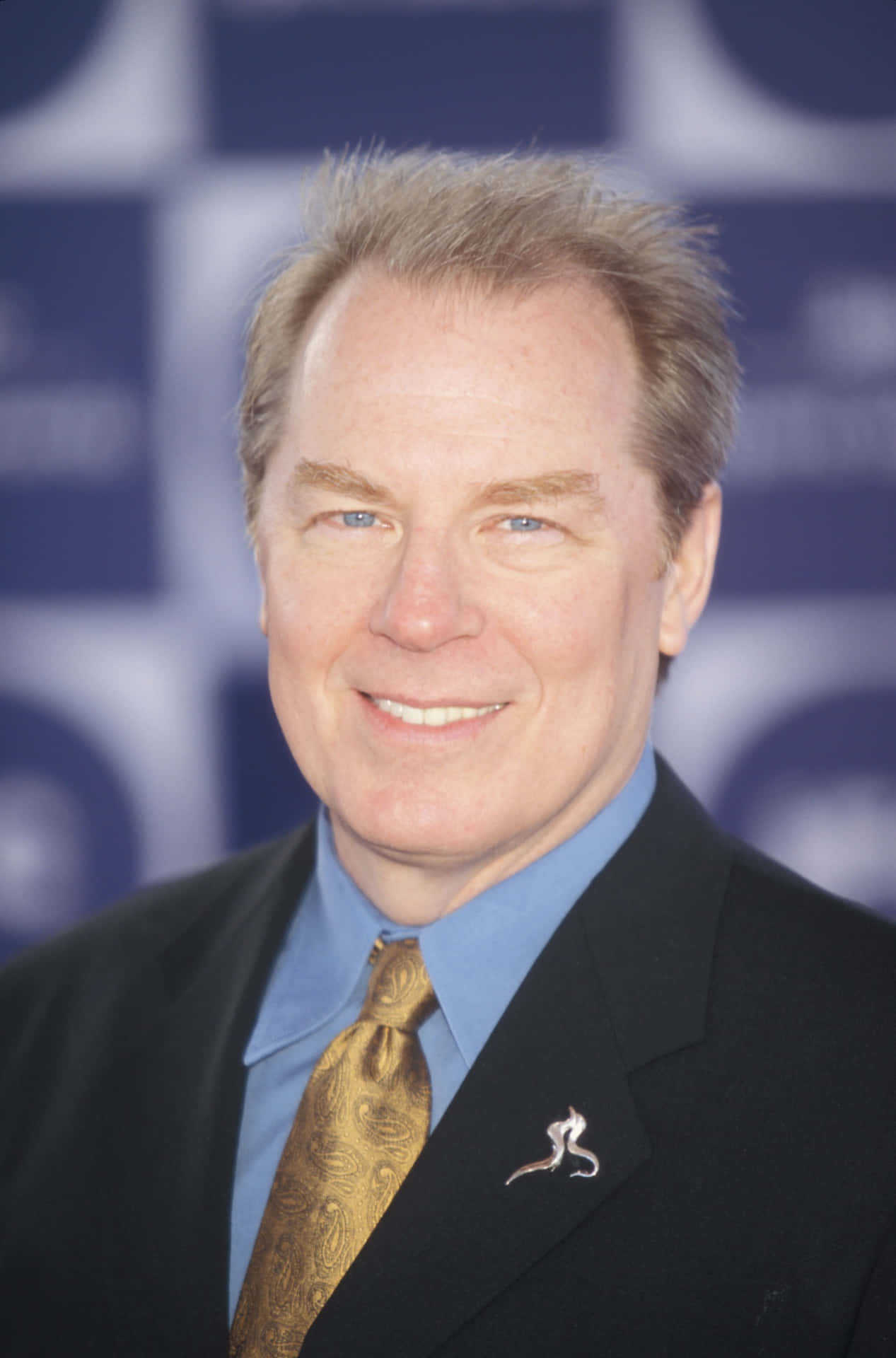 Michael Mckean 桌布 壁紙 — 一位微笑的男子身穿黑色西裝、藍色襯衫和金色佩斯利領帶，在模糊的藍白格仔背景前擺姿勢。