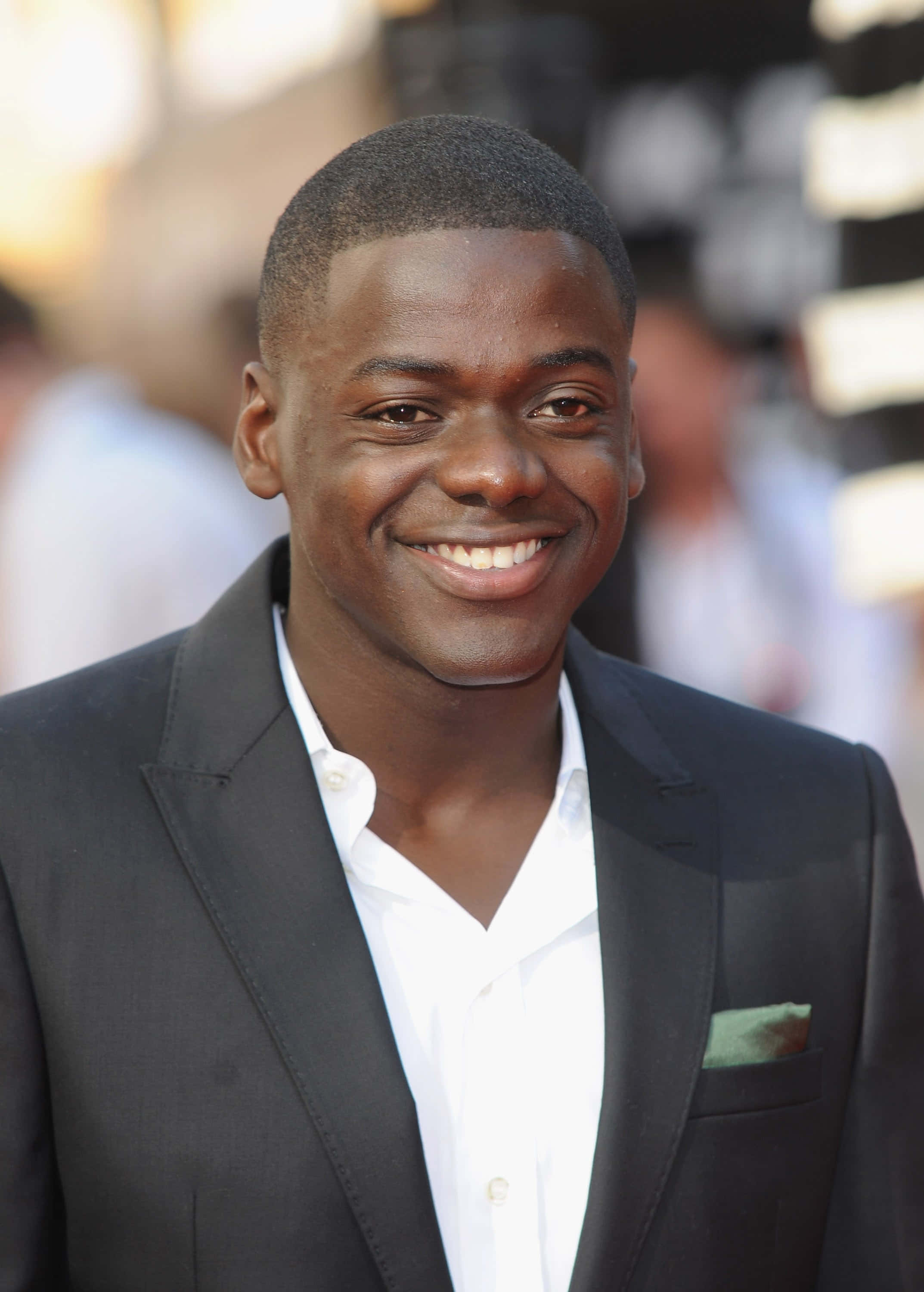 Daniel Kaluuya 桌布 壁紙 — 一位微笑的男子身穿黑色西裝和白色襯衫，在明亮的戶外活動中自信地擺姿勢，背景燈光模糊。